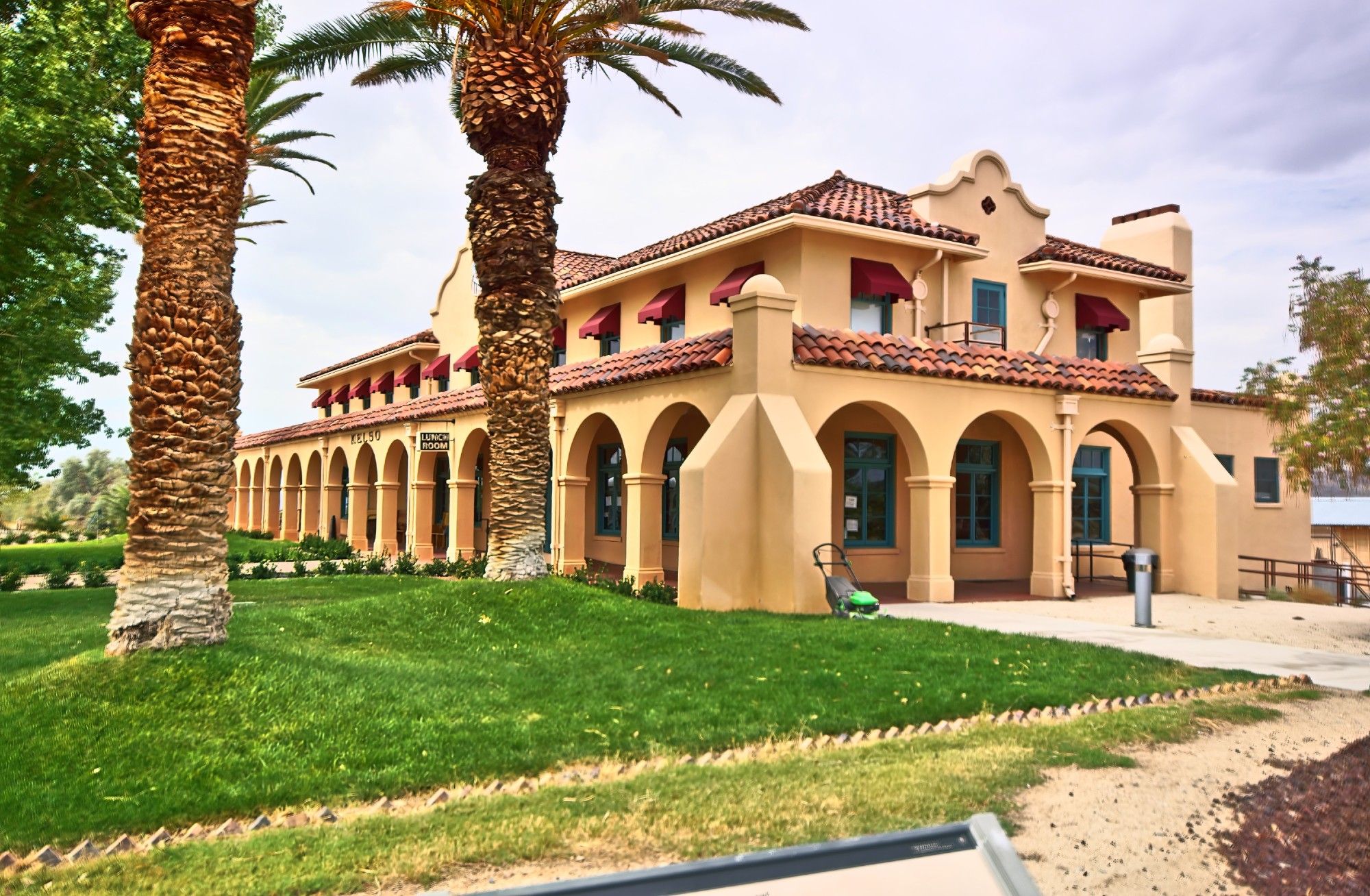 On the left, a large green lawn with two large palm trees, on the right a tan-coloured stucco building with a red tiled roof in mission revival style