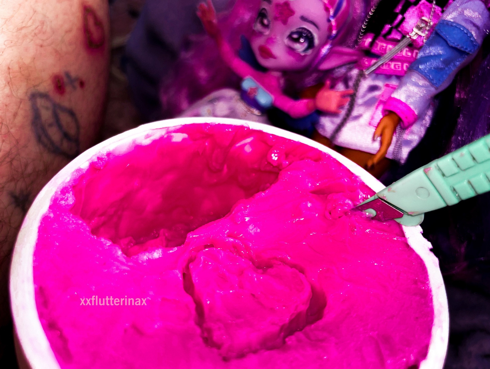 photo of a tub of hot pink hair dye with a heart shape carved into the dye. there is a disposable surgical scalpel sticking out of the dye after being used to carve the heart. there is a pink fairy fashion doll with her arms raised in the background, next to another fashion doll. angel’s leg is partially visible in the mid ground. his skin is pale, and he has a stick and poke tattoo of a UFO alongside a couple stars, an inflamed and scabbed over heart shape outline that has been recently been burnt into his skin, and a few pinprick burns among the space scene on his shin.