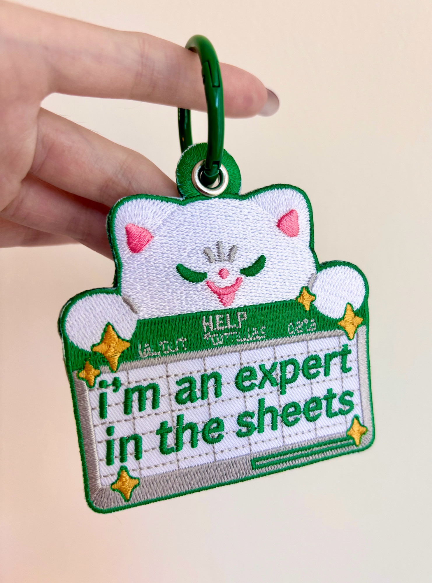 A photo of an embroidered keychain with a green metal key ring. The embroidery is of a smug looking cartoon cat holding an excel spreadsheet that reads “I’m an expert in the sheets”  