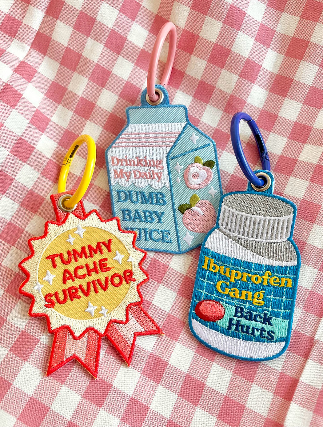 A picture of three embroidered keychains laying on pink gingham fabric. The one on the left is red and yellow and is shaped like an award ribbon and reads “TUMMY ACHE SURVIVOR”. The one in the middle is of a light blue juice carton that reads “drinking my daily DUMB BABY JUICE” and has peaches on it. The one on the right is shaped like a medicine bottle and reads “ibuprofen gang back hurts”