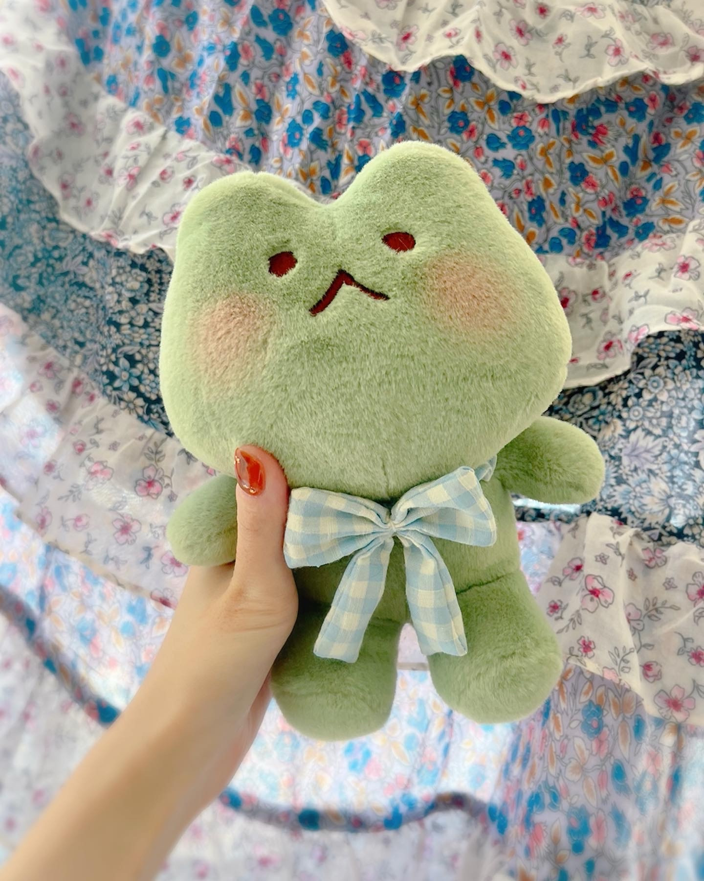 Photo shows a hand holding a frog plush. The plush is light green and has a light blue gingham bow around its neck 