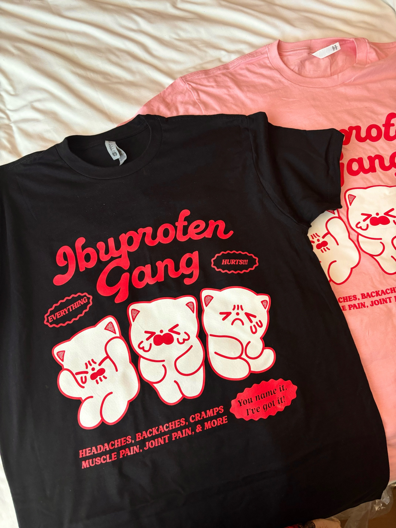 Picture of two tshirts. The artwork on the shirts reads “ibuprofen gang” and shows three cats- one on the left is clutching its head in pain, one in the middle is crying and holding its tummy, and the one on the right is also in pain holding its tummy. The rest of the shirt reads “headaches, backaches, cramps, muscle pain, joint pain & more. You name it, I’ve got it!”