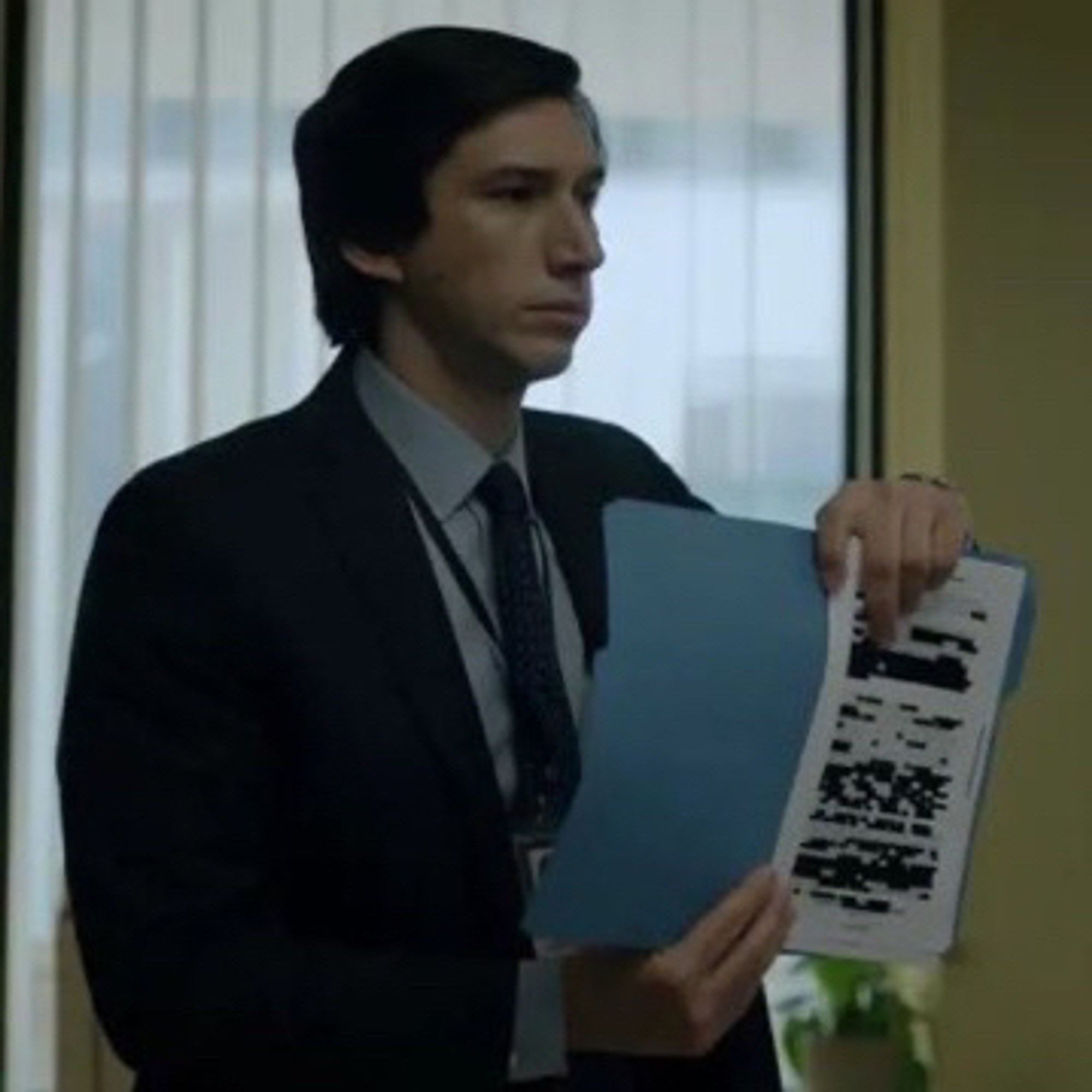 Adam driver wearing a black suit and tie and grey collared shirt while holding up a redacted file in an office.