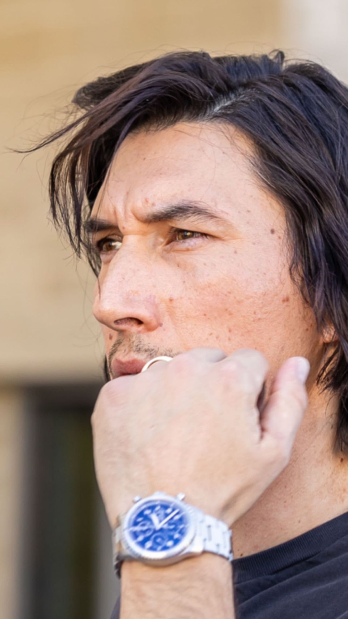 Adam driver with wind swept hair, goatee stubble, and a silver ring and watch on his hand in a fist on his face. He’s wearing a black t shirt