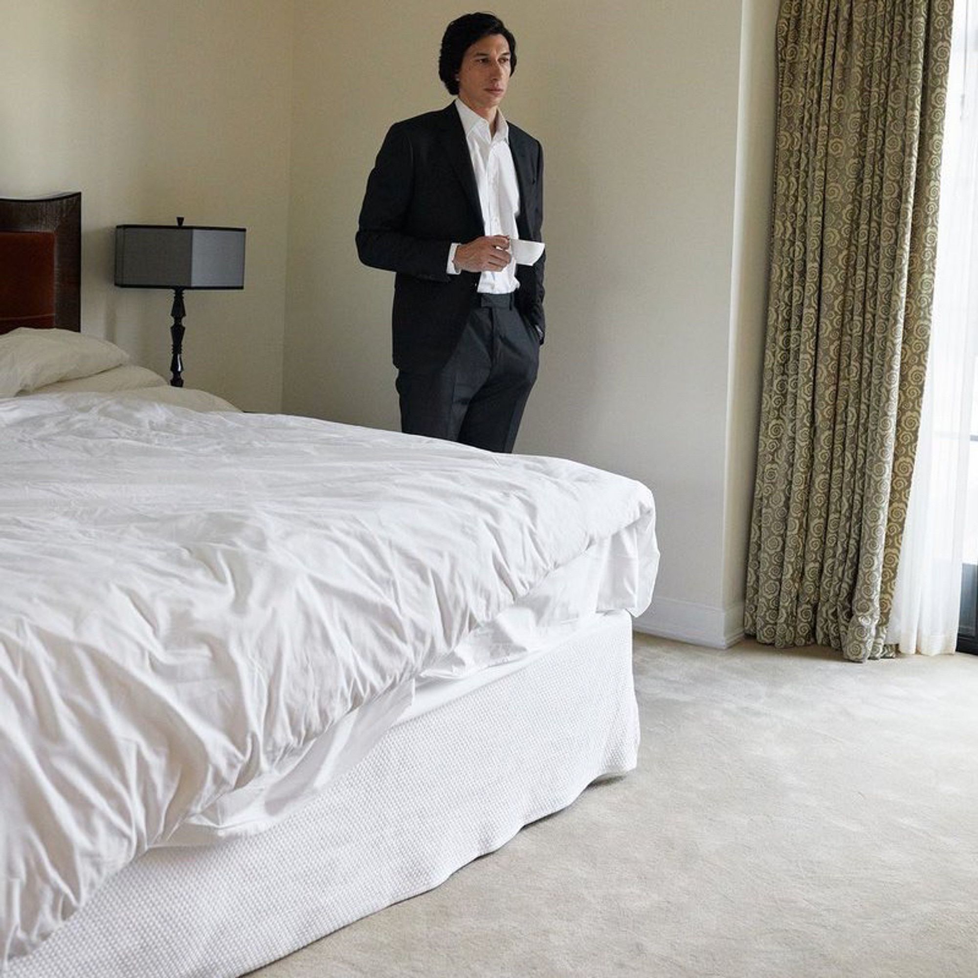 Adam driver with one hand in his pocket wearing a black suit and white collared shirt while holding a tea cup as he stands next to a white bed and grey lamp and green curtains in a hotel room