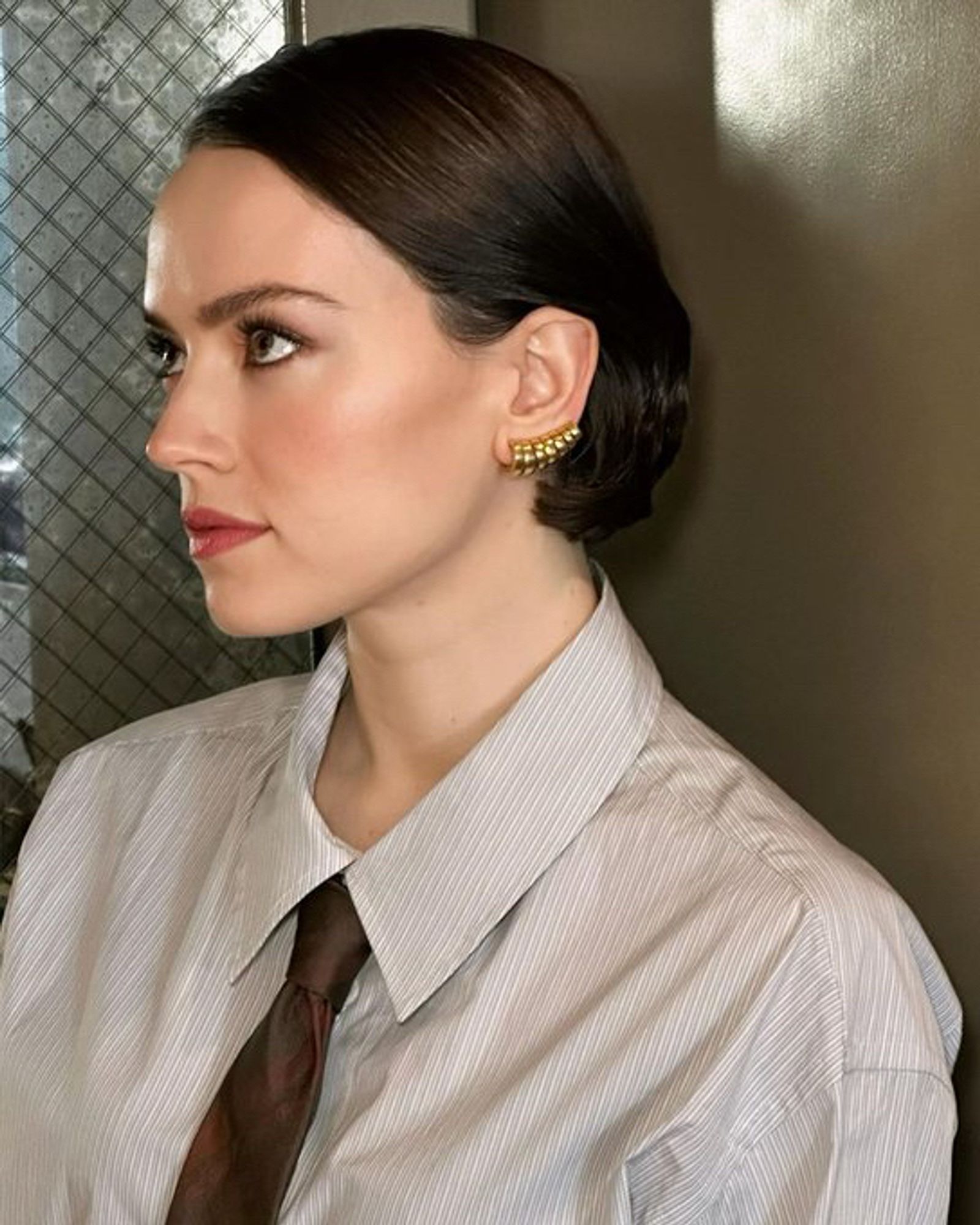 Daisy Ridley with hair slickly parted to side, wearing gold multihooped earrings, striped grey collared shirt and brown tie as she looks up and stands next to a glass netted patterned window door