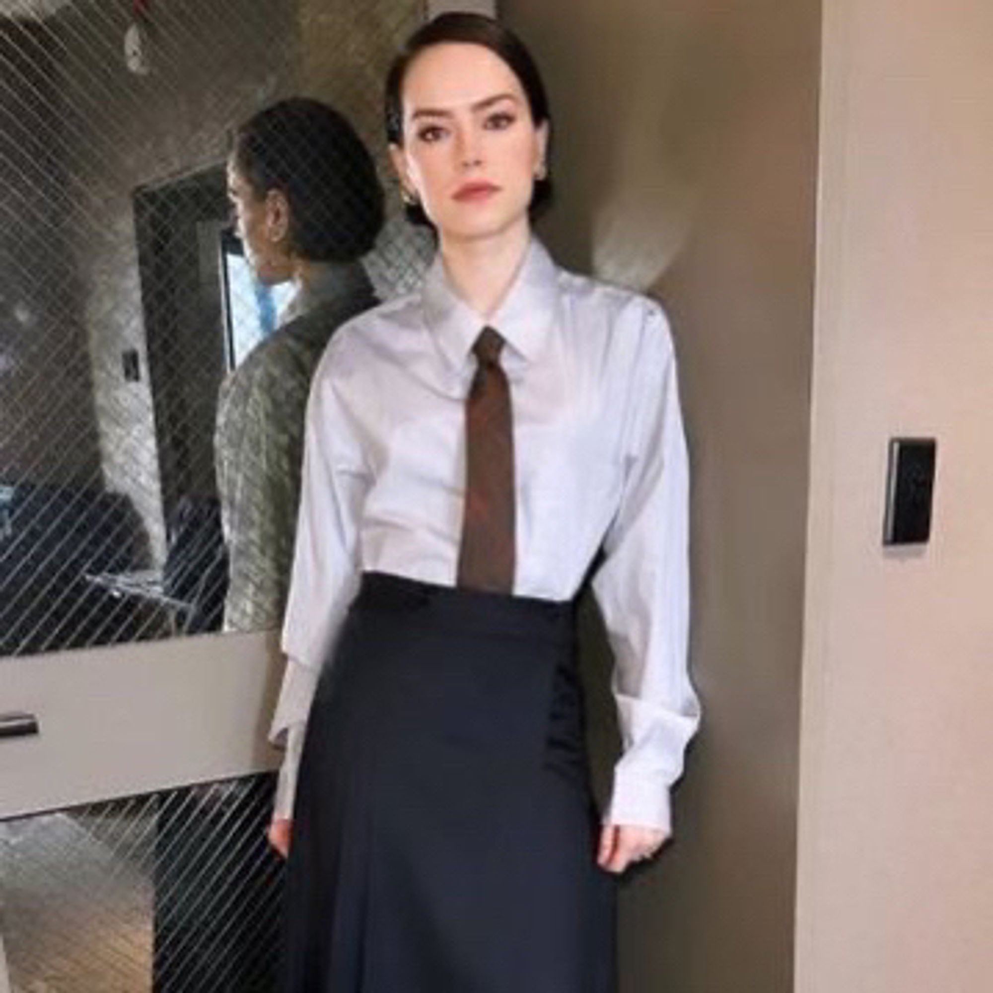 Daisy Ridley wearing hair short and slicked to the side, gold multihoop earrings, grey or white collared shirt, brown tie and black long skirt. She’s posed next to a glass door