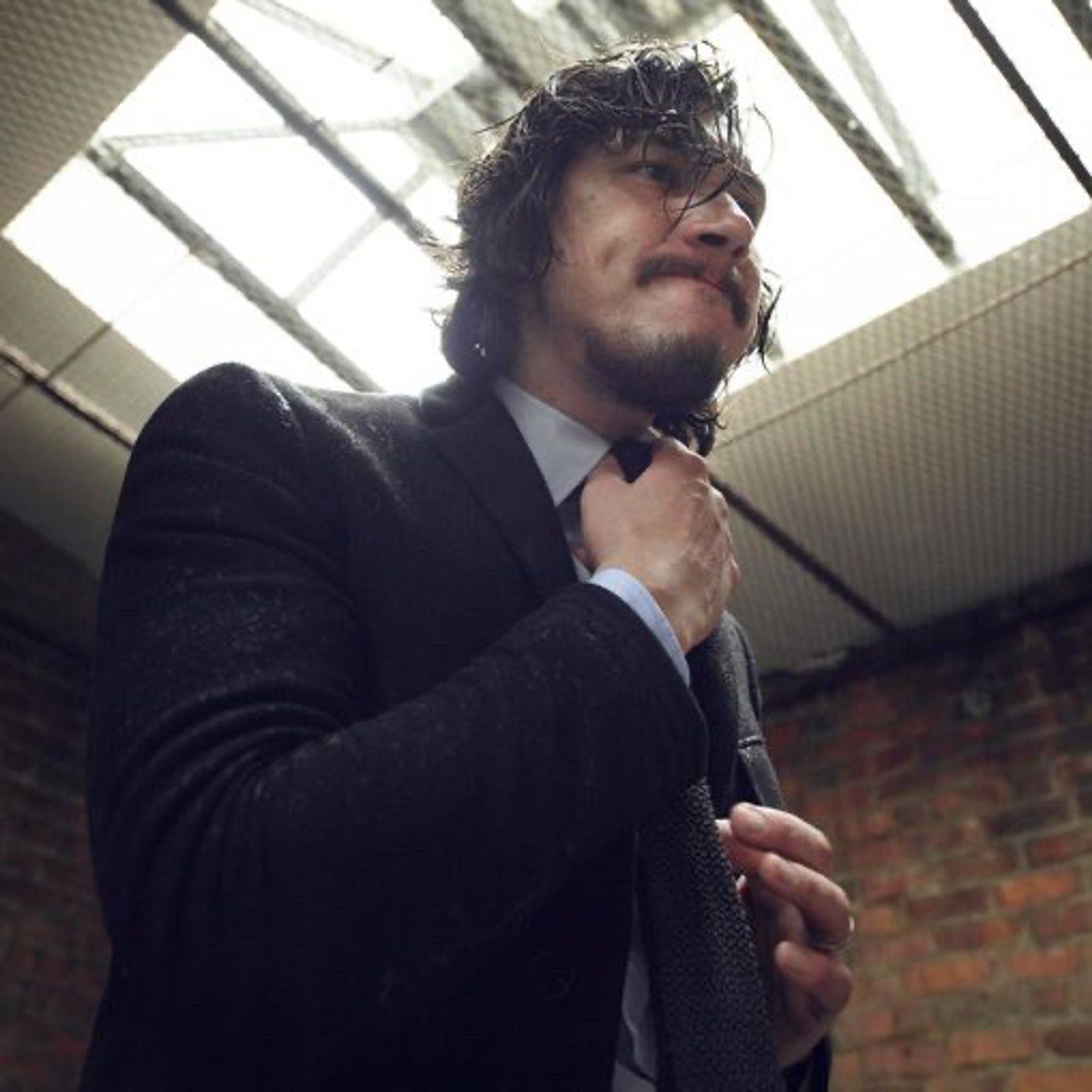 Adam driver wearing a wet black silk suit and tie and grey collared shirt and has his hands adjusting his tie with his hair wet and shaggy and with a goatee as he stands in a brick walled room with a grate above between the room and the skylight