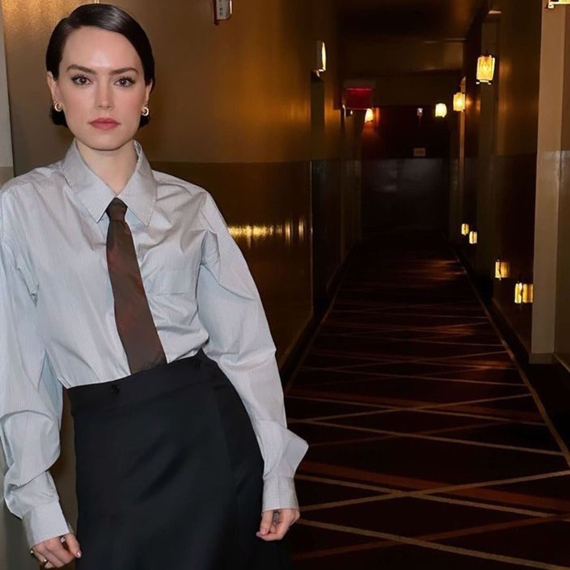 Daisy Ridley wearing multihooped gold earrings, tucked in grey collared shirt, brown flannel patterned tie, black long skirt and hair slicked and parted to the side while leaning on a wall of a hotel with light sconces on the bottom of the walls and dark burgundy yellow and orange carpet