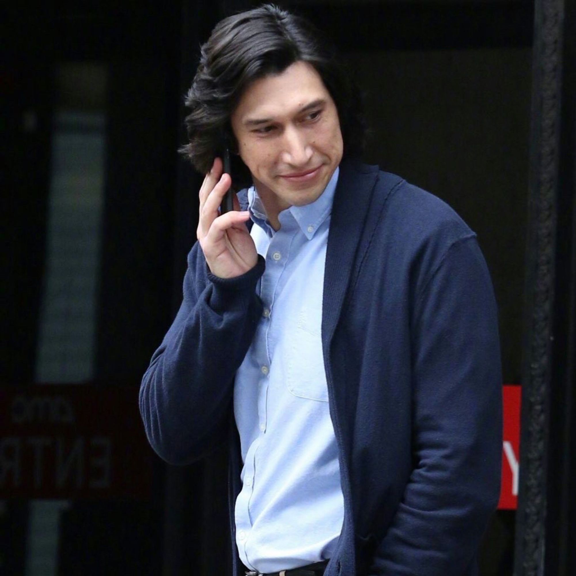 Adam driver as Charlie in marriage story smiling as he has a cellphone to his ear wearing a blue cardigan and light blue collared shirt