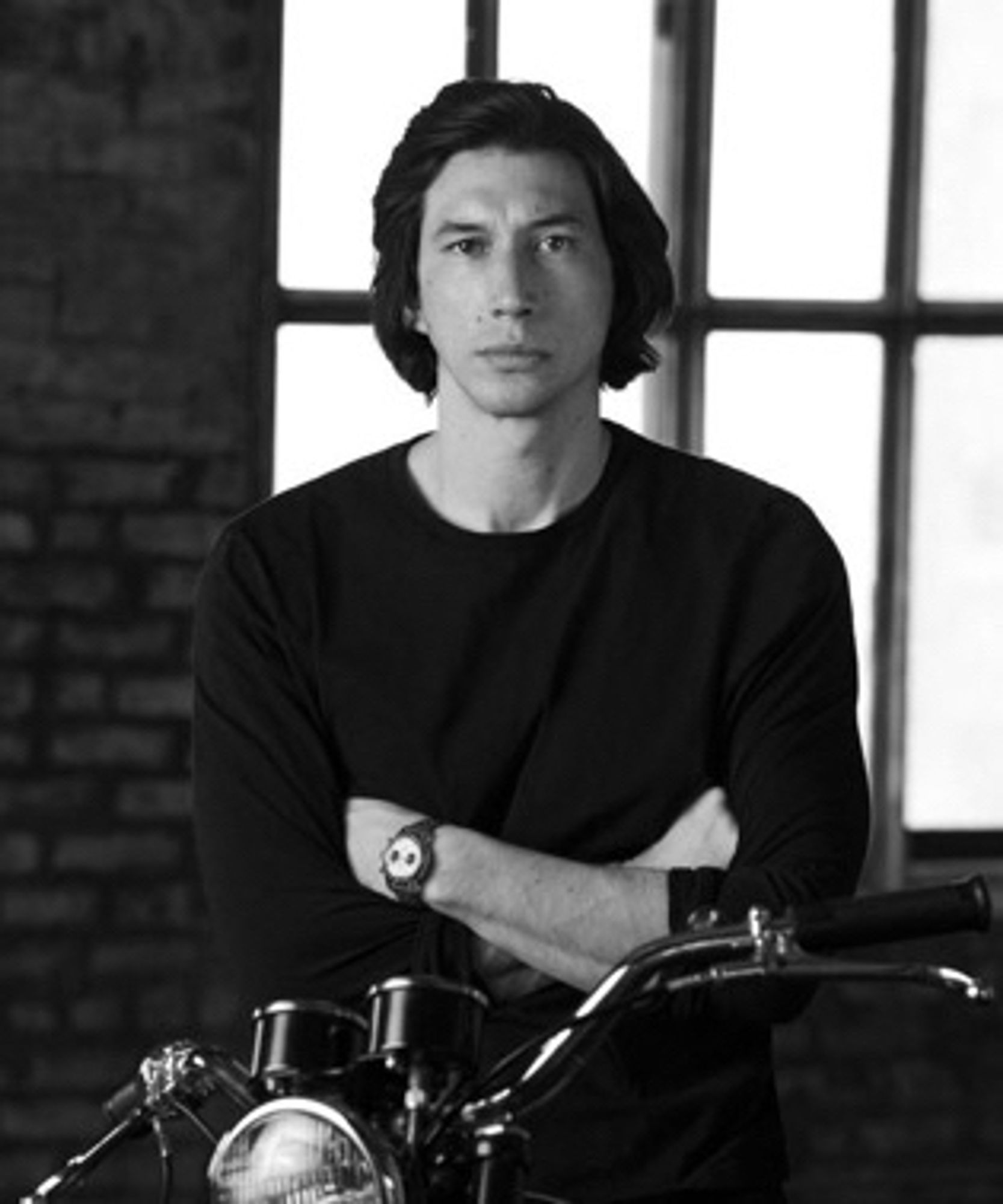 Adam driver on a motorcycle with his arms crossed, wearing a black shirt, Breitling watch, and a brick wall and window behind him