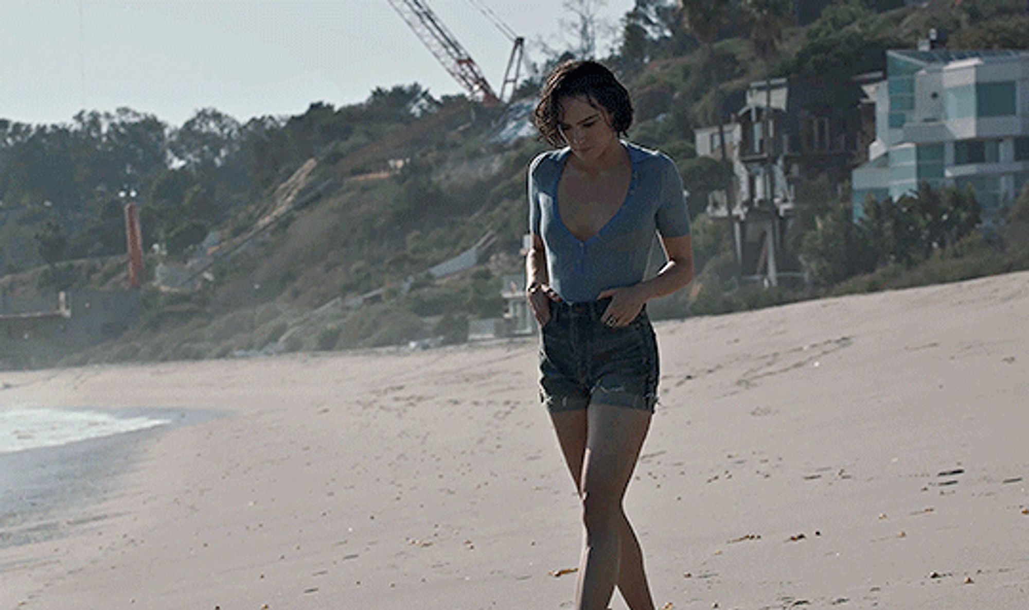 Daisy Ridley,hands in her pockets, walking on the beach, homes and cranes on the hillside behind her, soft waves going up the sand, wearing denim shorts with the cuffs folded up& a light blue short sleeved top with scoop neck. Her hair is short, Bob length and wet and wavy