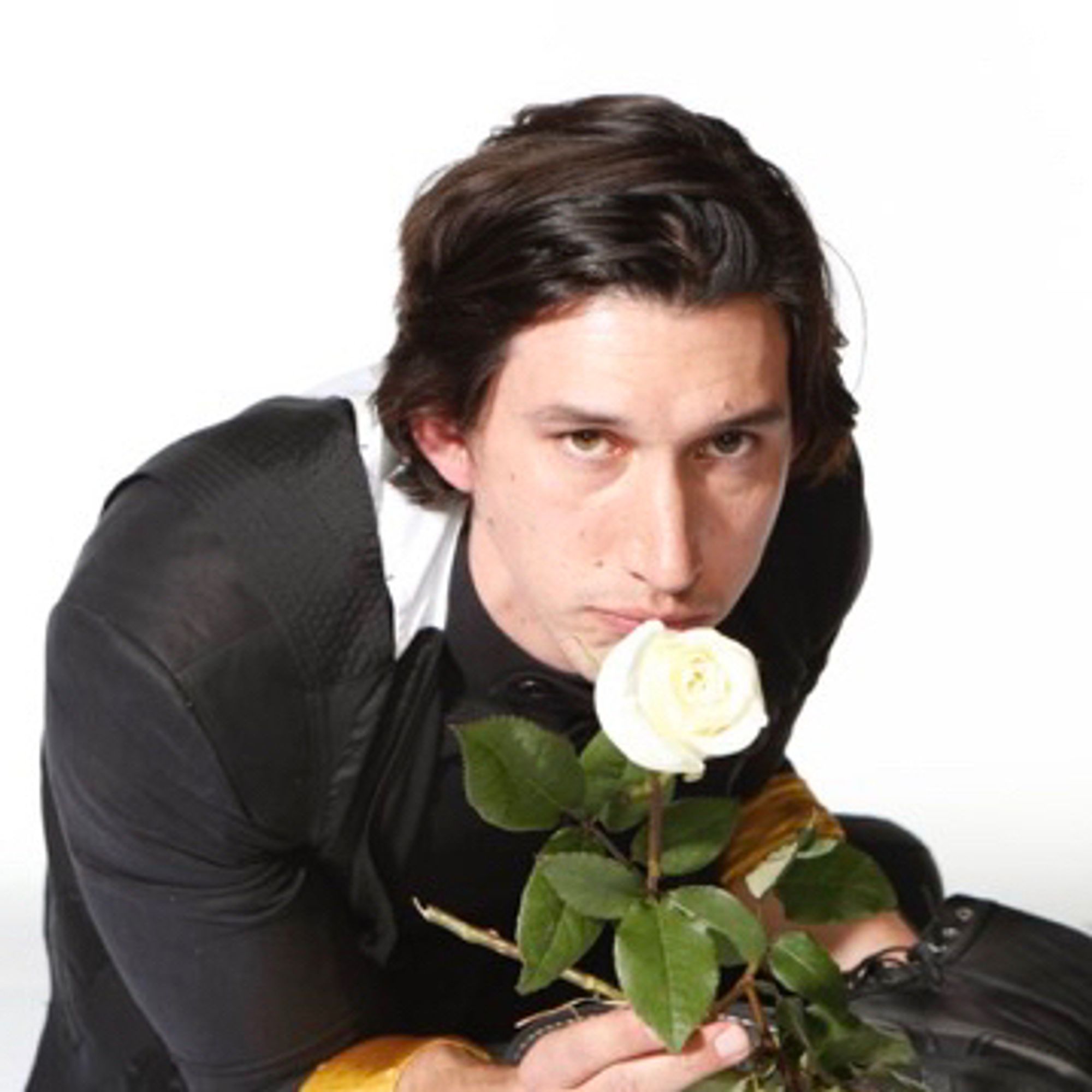 Adam driver wearing a black suit and holding a boot on his lap and a white rose pressed to his lips