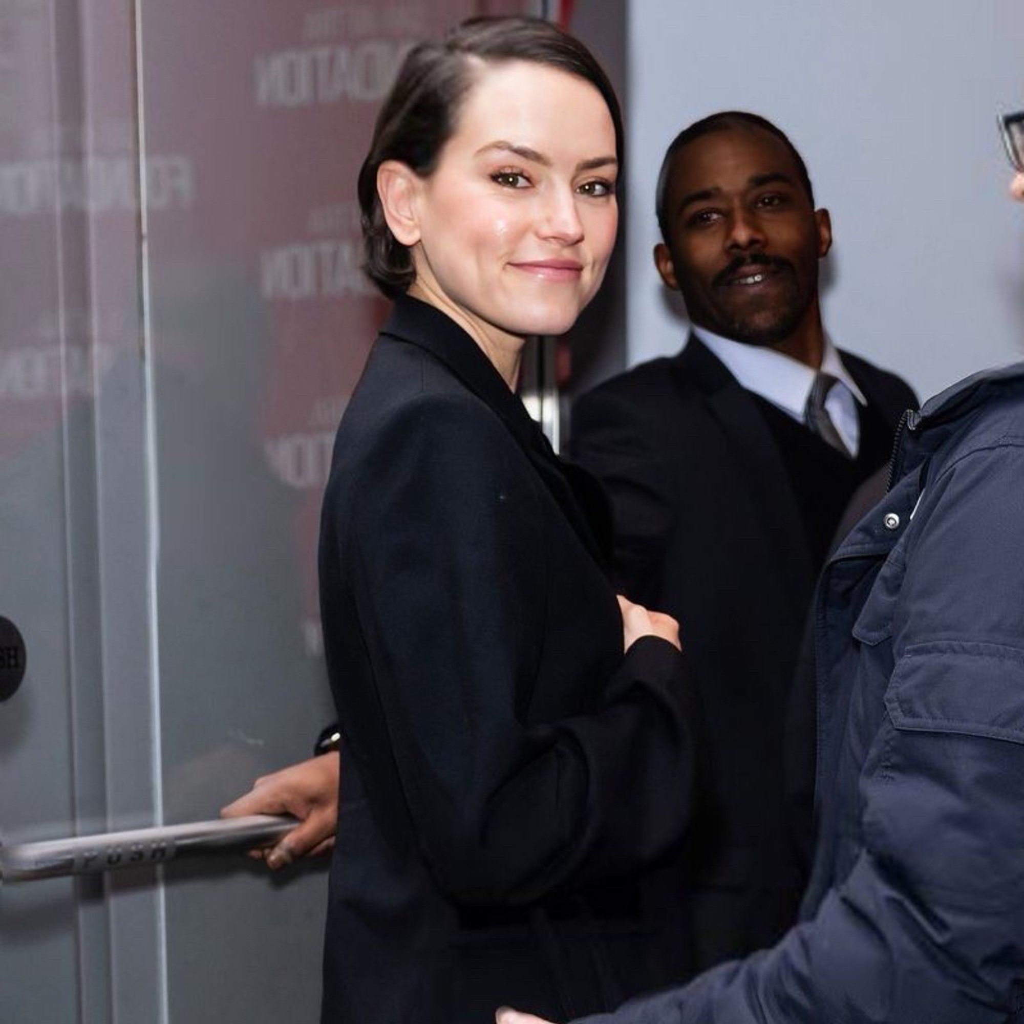 Daisy Ridley smiling, short hair parted to side and wearing a black blazer and two people near her as a door is being held open for her, a wall with the words “sag aftra foundation” is seen in the reflection