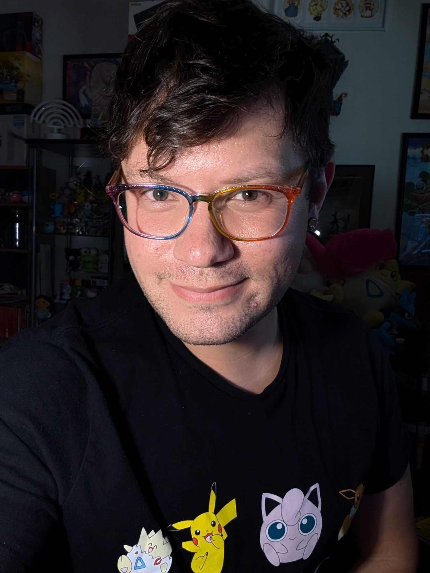 SaskGaymer smiling. He's wearing a black shirt with pokemon on it and rainbow framed glasses. He has dark brown hair. Pokemon plushies and various collectibles in the background.