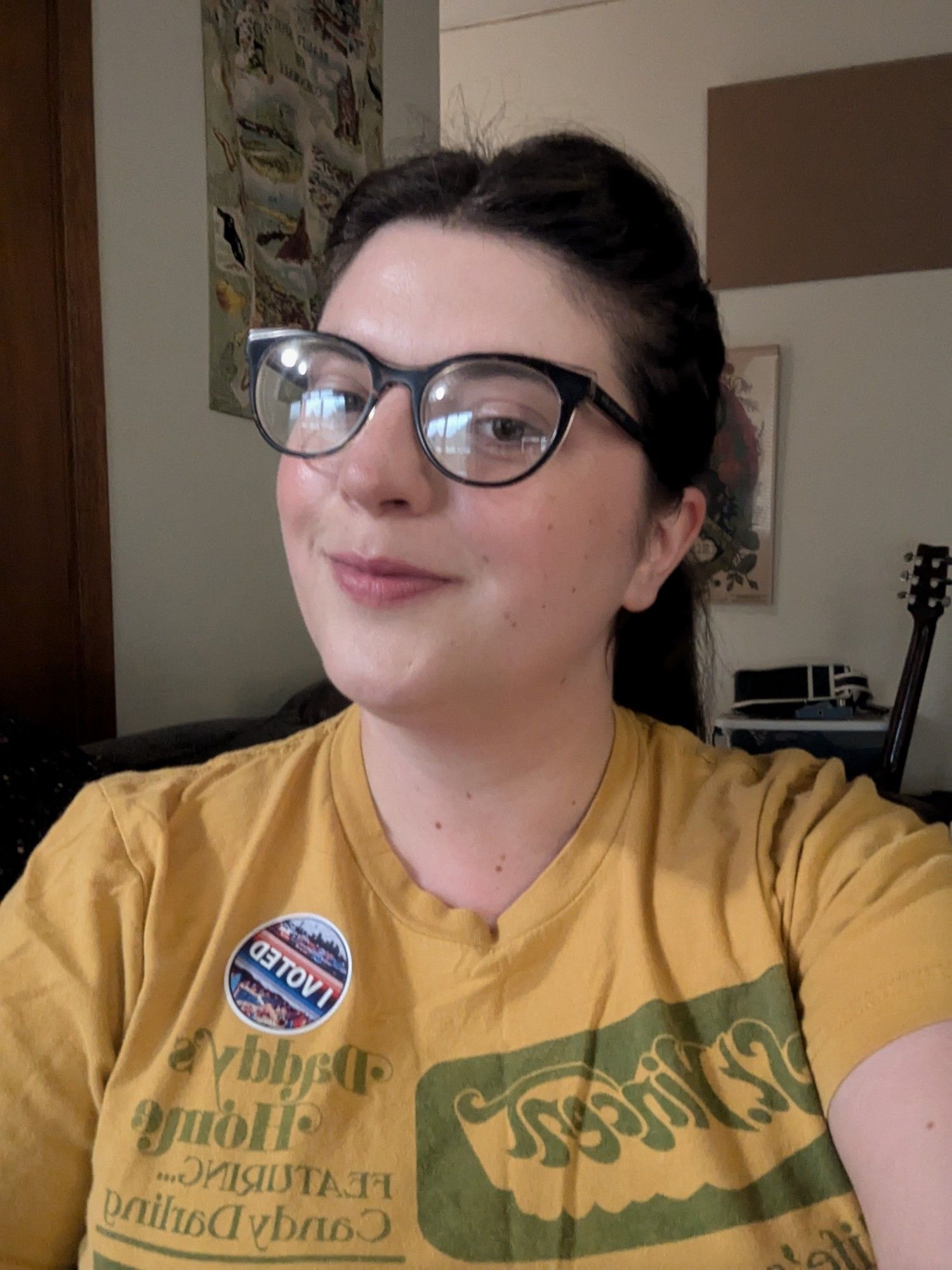 Brunette woman wearing glasses and a yellow St Vincent tee. Her I Voted sticker slapped on the left side of her chest.