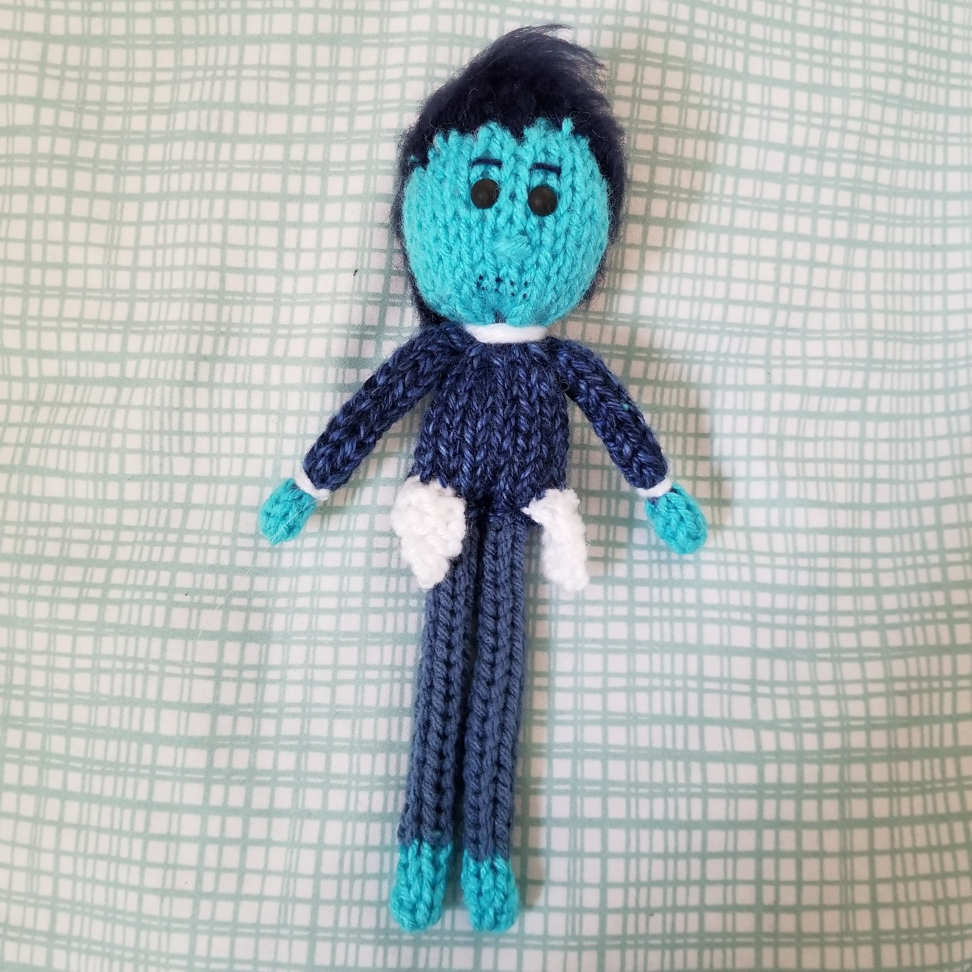 A knit doll of Percy Reed from Hello from the Hallowoods. His entire pallette is blue to attempt to show his ghost nature and he has short hair, matte black eyes, and small needle holes around his mouth. He is wearing a collared shirt, sweater, and pants (season 2 wardrobe).