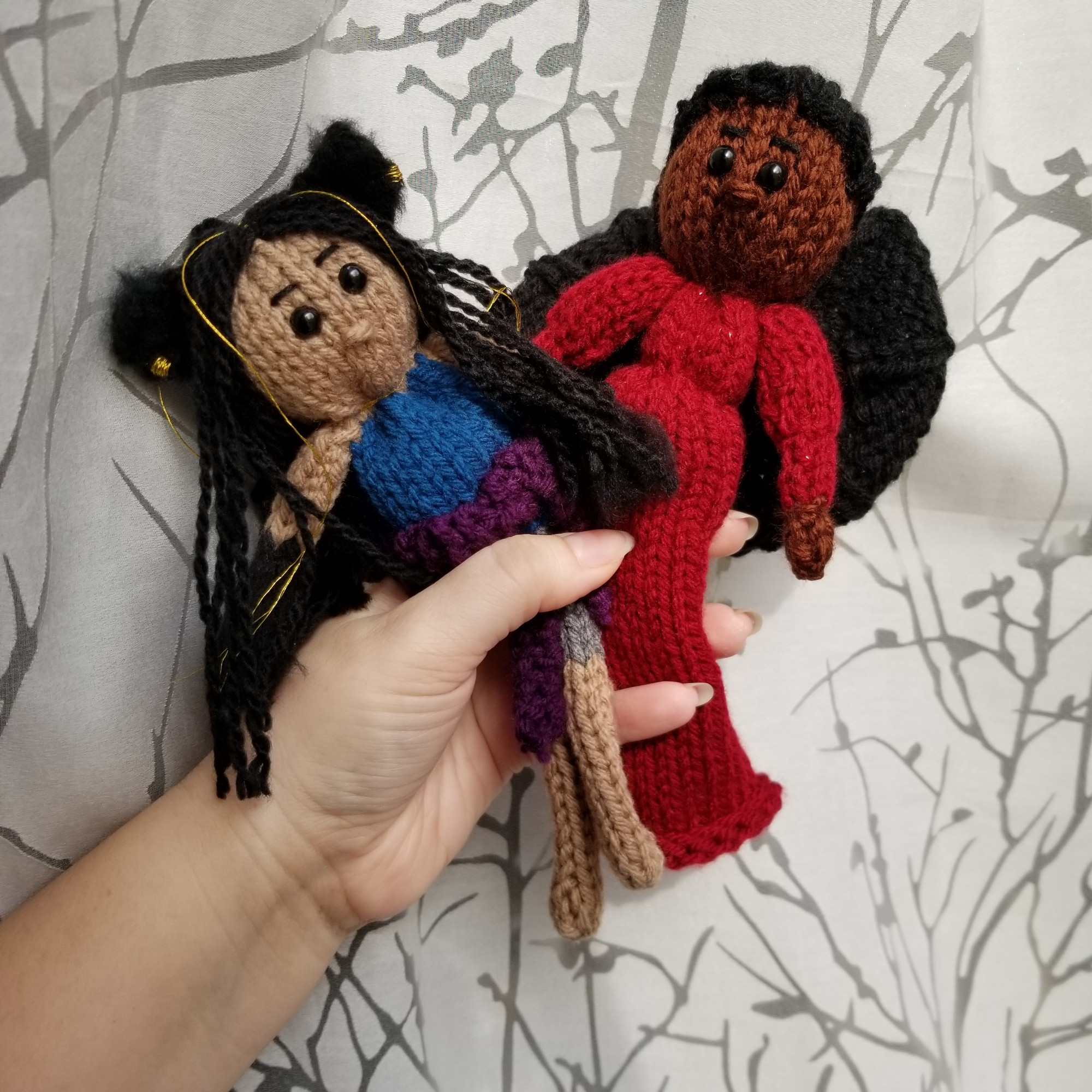 Two knit dolls of Yaretzi and The Countess from Hello from the Hallowoods. Yaretzi is tan-skinned with long black hair, peppered with gold thread. She's wearing a sleeveless blue top and grey capris with a purple sweatshirt wrapped around her waist. On her head are fluffy black wolf ears with gold earrings and halfway up her arms is black fur starting from her hands. The Countess is Black with short black hair, wearing a red, form fitting dress with puffy sleeves.On her back are two black bat wings.