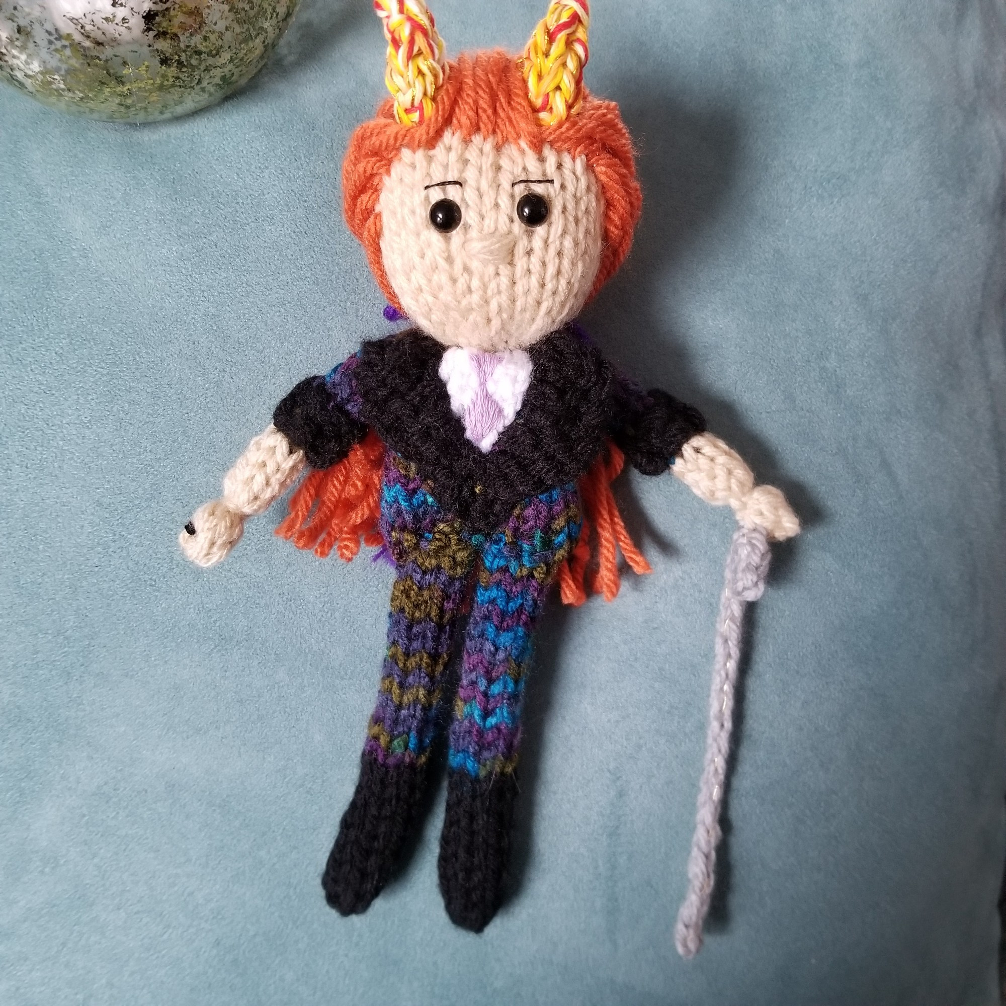 A solo photo of the Polly doll. Polly is pale-skinned and is wearing a colorful suit. His orange hair is tied back, and he carries a cane. On his head are two fiery horns. He wears a black ace ring on his right hand.