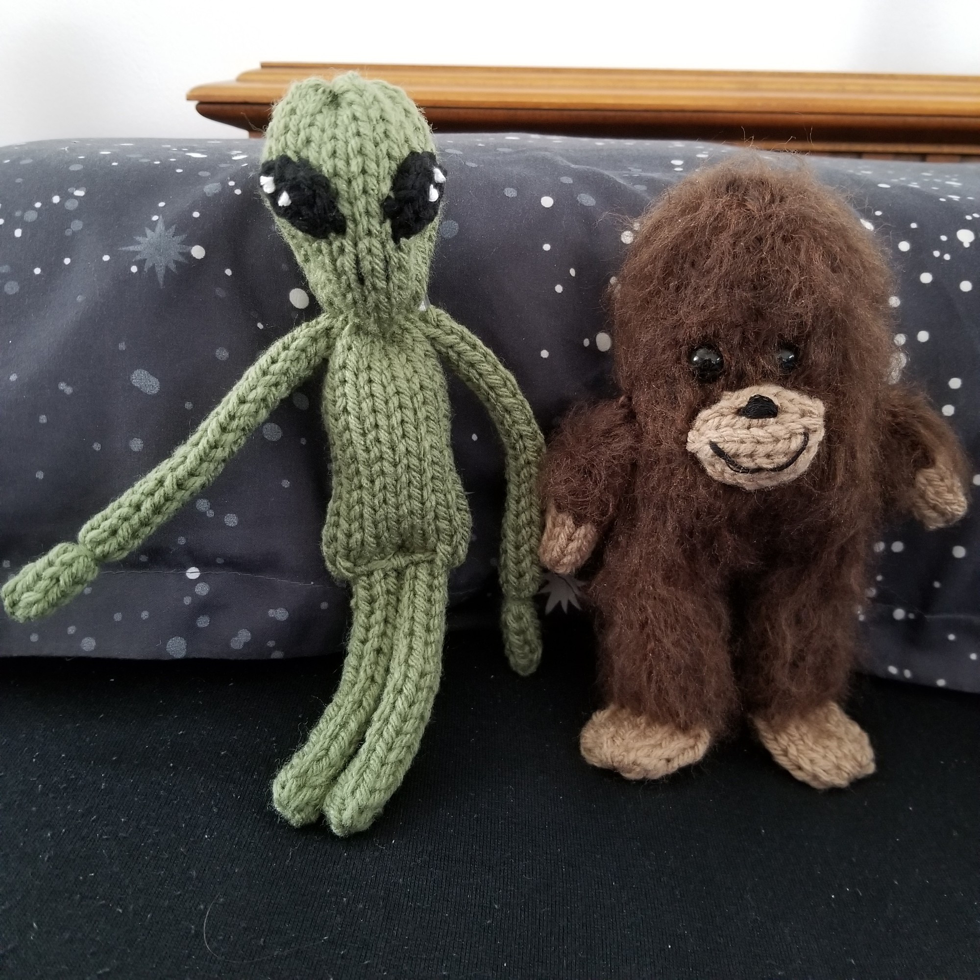 Two knit dolls of an alien and a bigfoot. The alien is light green with a body and arms longer than its legs, and big almond shaped eyes. The bigfoot is shorter than the alien. Fuzzy brown fur covers its whole body except the light brown hands, feet, and muzzle.