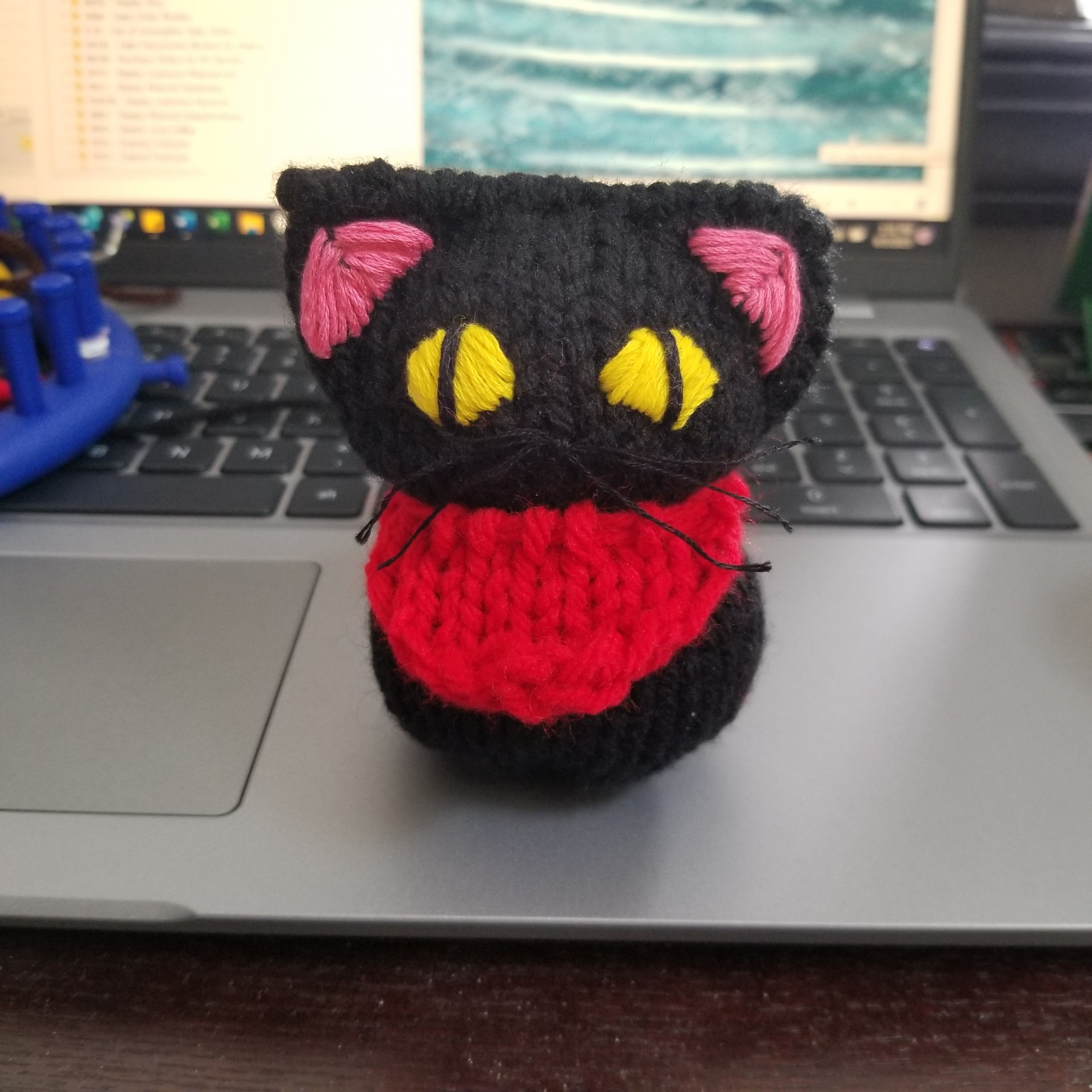 A knit doll of a small black cat with yellow eyes and a red bandana.