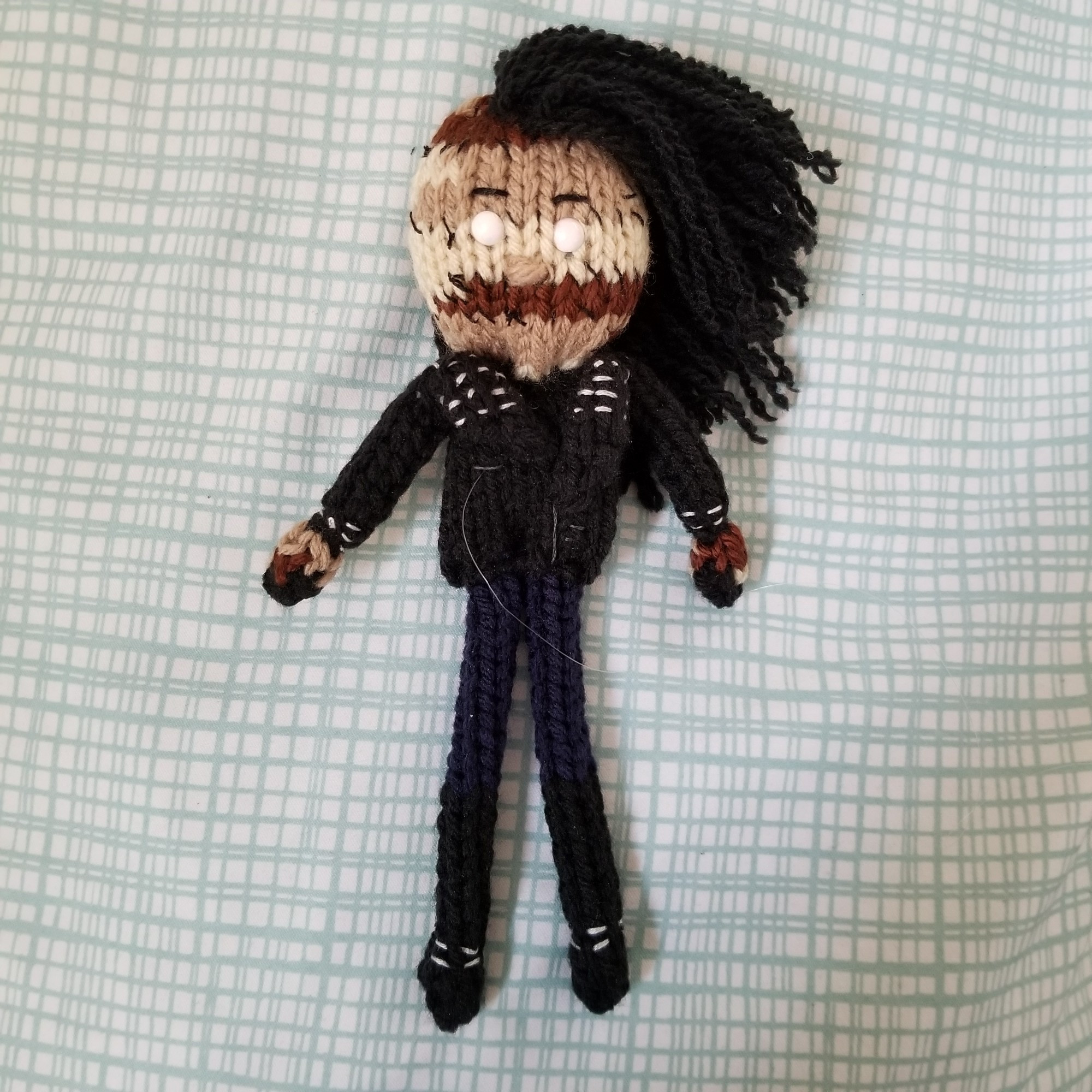 Knit doll of Diggory Graves from Hello from the Hallowoods. Diggory has multiple skin tones stitched together on their face and hands, and black hair in an unstyled mohawk. They're wearing a black leather jacket, dark blue pants, and tall black boots. Their eyes are painted white.