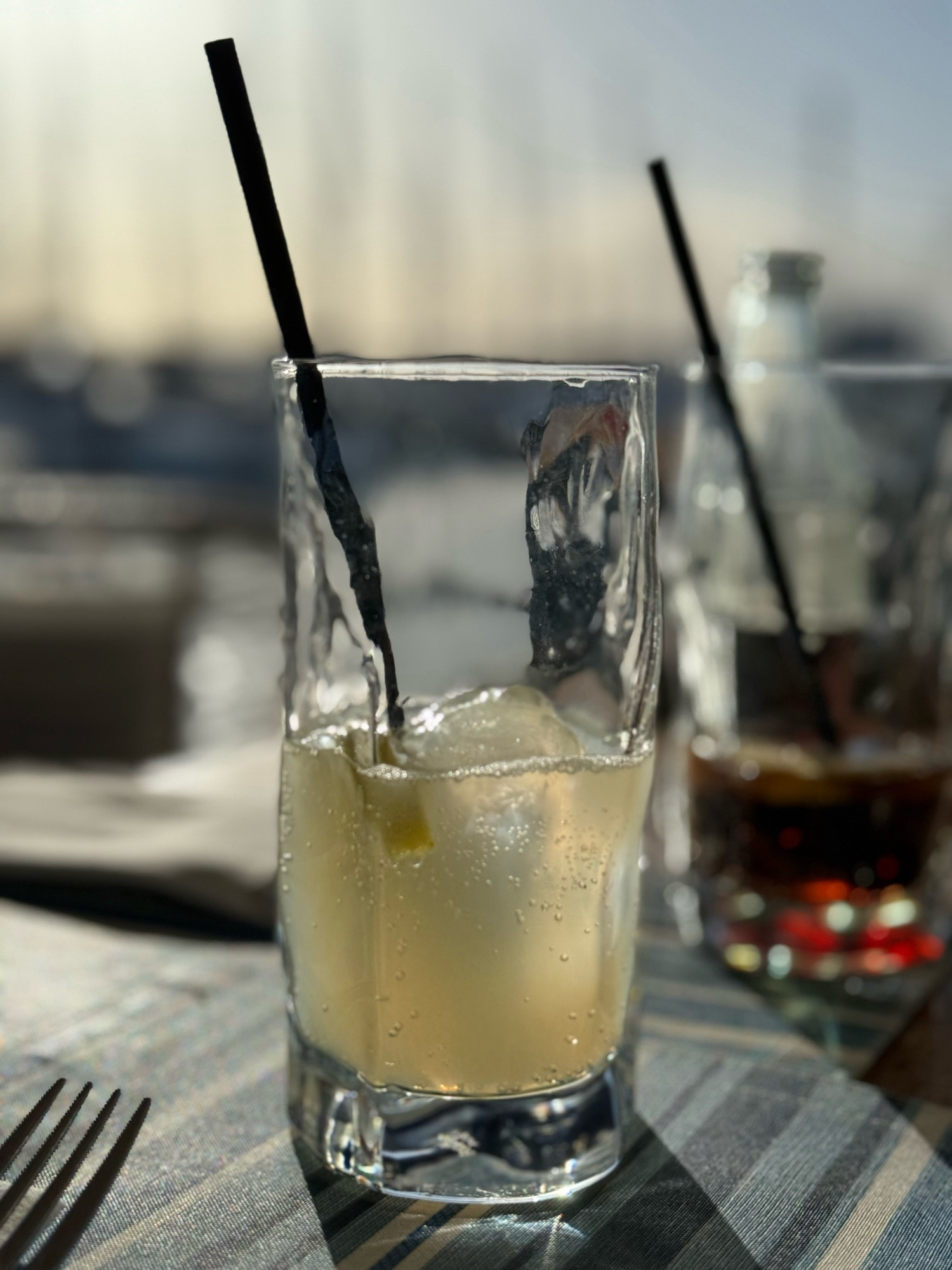 Ein Glas Limonade auf einem Tisch in der Sonne. Im Hintergrund sind Maste von Segelbooten in einem Hafen verschwommen angedeutet.