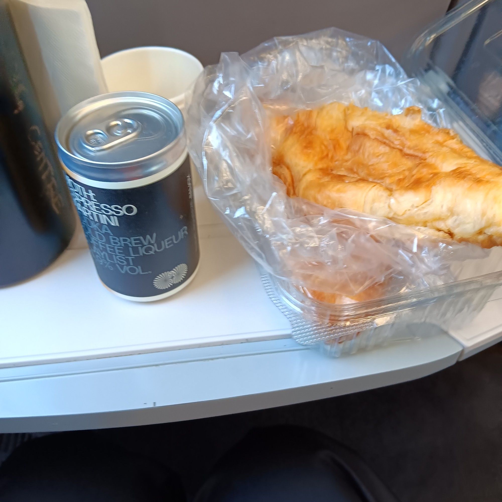 A fold down table on a train, with a water bottle, a tinned espresso martini and some croissants on it.