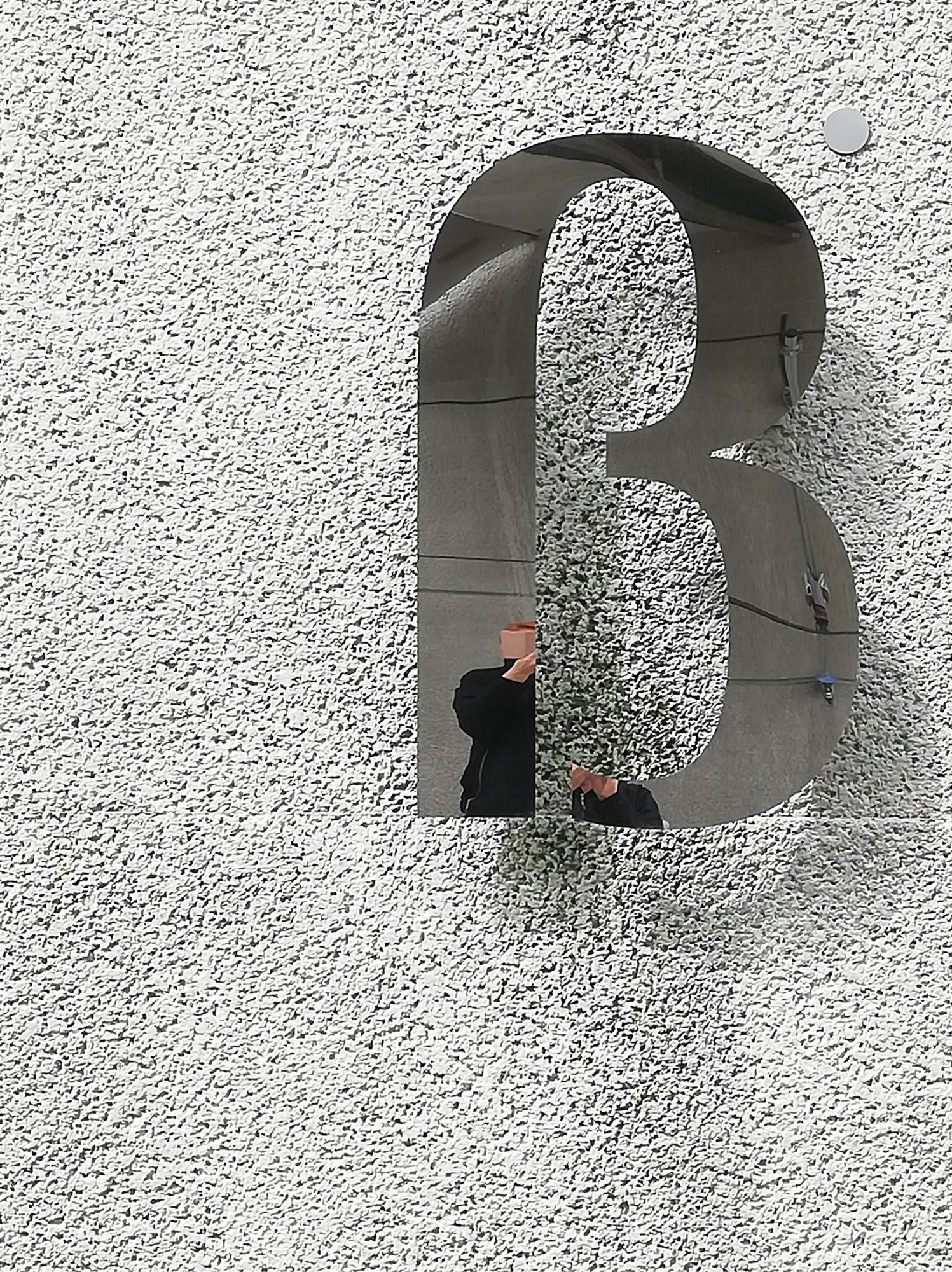 Der Buchstabe ß an einer Wand hängend, darin eine Spiegelung der Fotografierenden - Hand, teilweise der Oberkörper