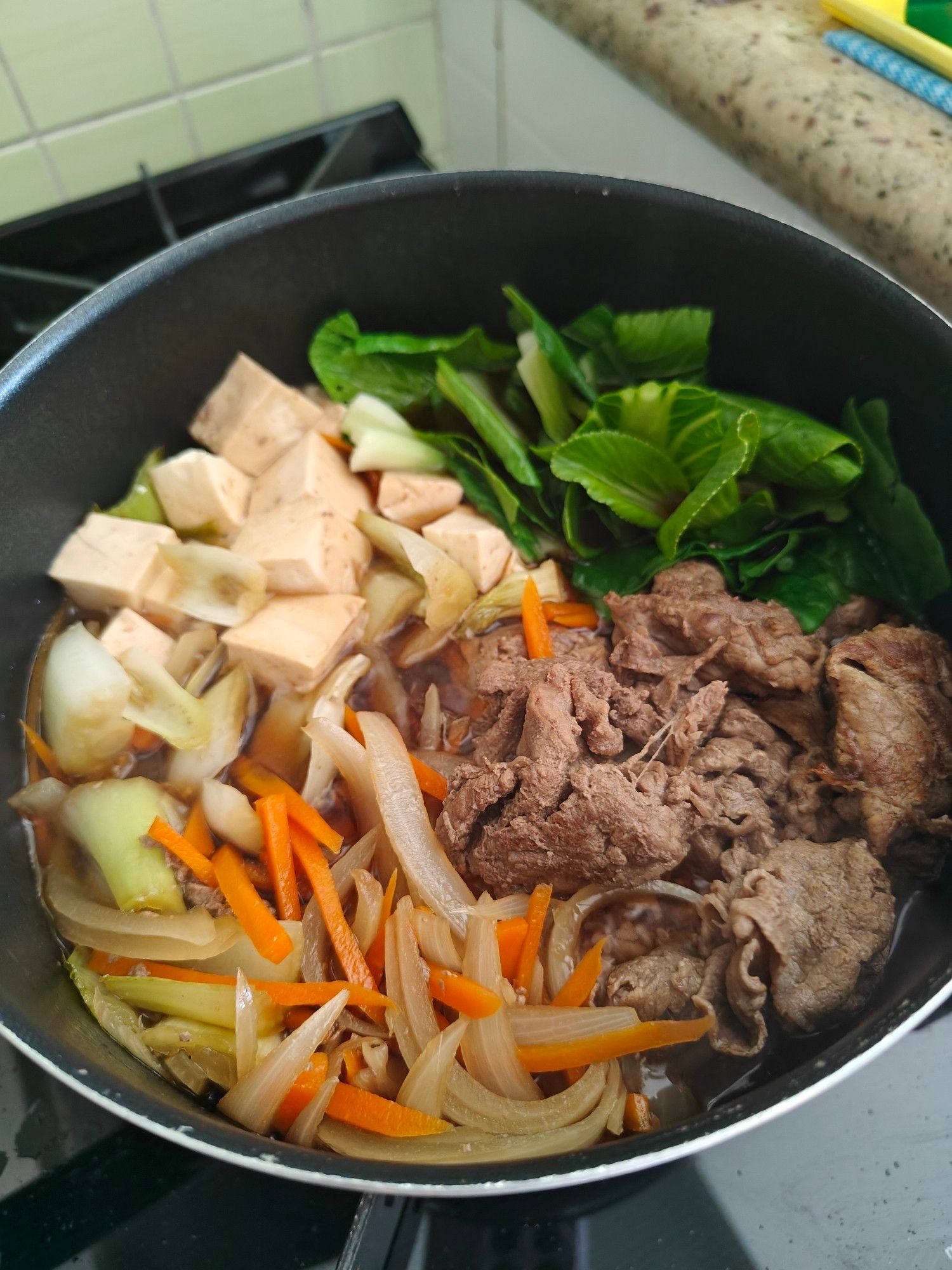 Panela com seções de carne, cebola e cenoura finas, talos de acelga, tofu em cubos e folhas verdes de acelga e espinafre.