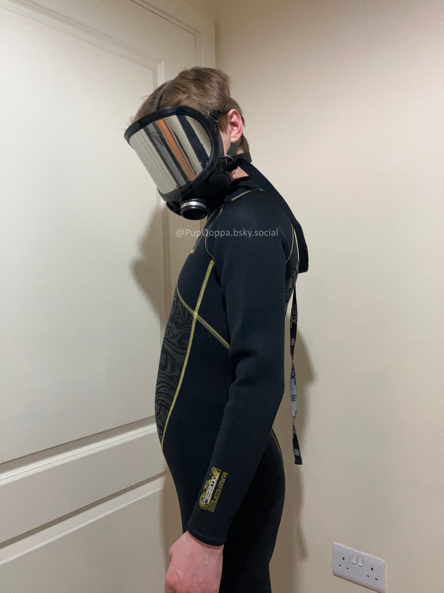 Picture of Qoppa in a neoprene wetsuit while wearing a mirrored gas-mask. It is in profile and looking sideways at the camera.