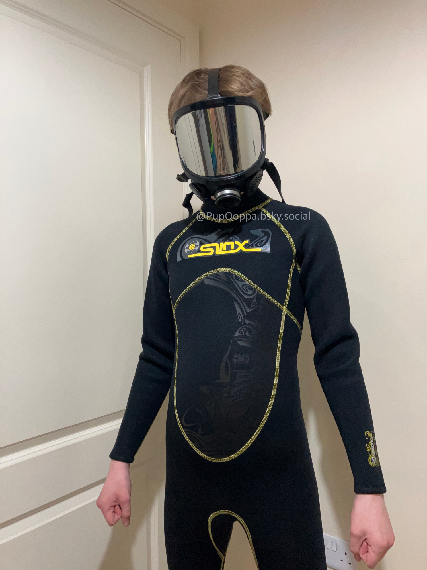 Picture of Qoppa in a neoprene wetsuit while wearing a mirrored gas-mask. It is standing straight in front of the camera.