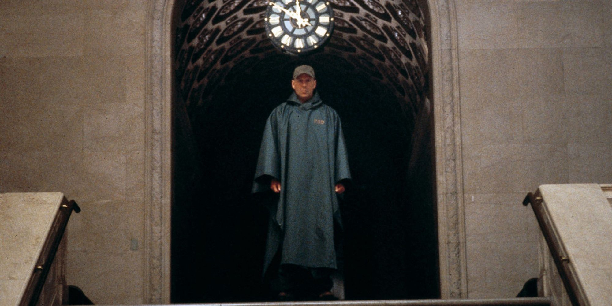 Bruce Willis (David) stands atop a grand flight of stairs in a train station. The camera looking towards him from a distant point at the base of said stairs. His dark green, rain poncho and hat dripping from the storm outside. A ornate clock sits behind him in the walkway he just came through. It is 12.50.