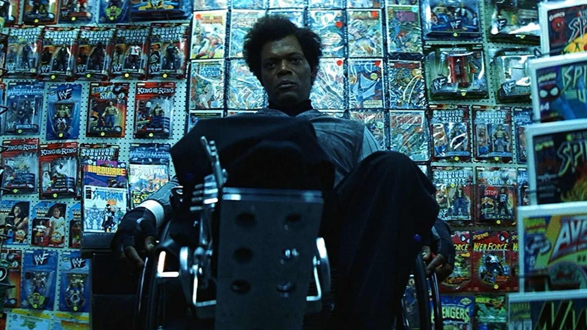 Samuel L Jackson (Mr Glass) sits in his wheelchair at the back of a comic book store. The camera viewing him from the front, low, looking up towards him. The wall behind clad in comics and toys of all kinds. Mr Glass is lost in thought. Completely gripped by his current predicament