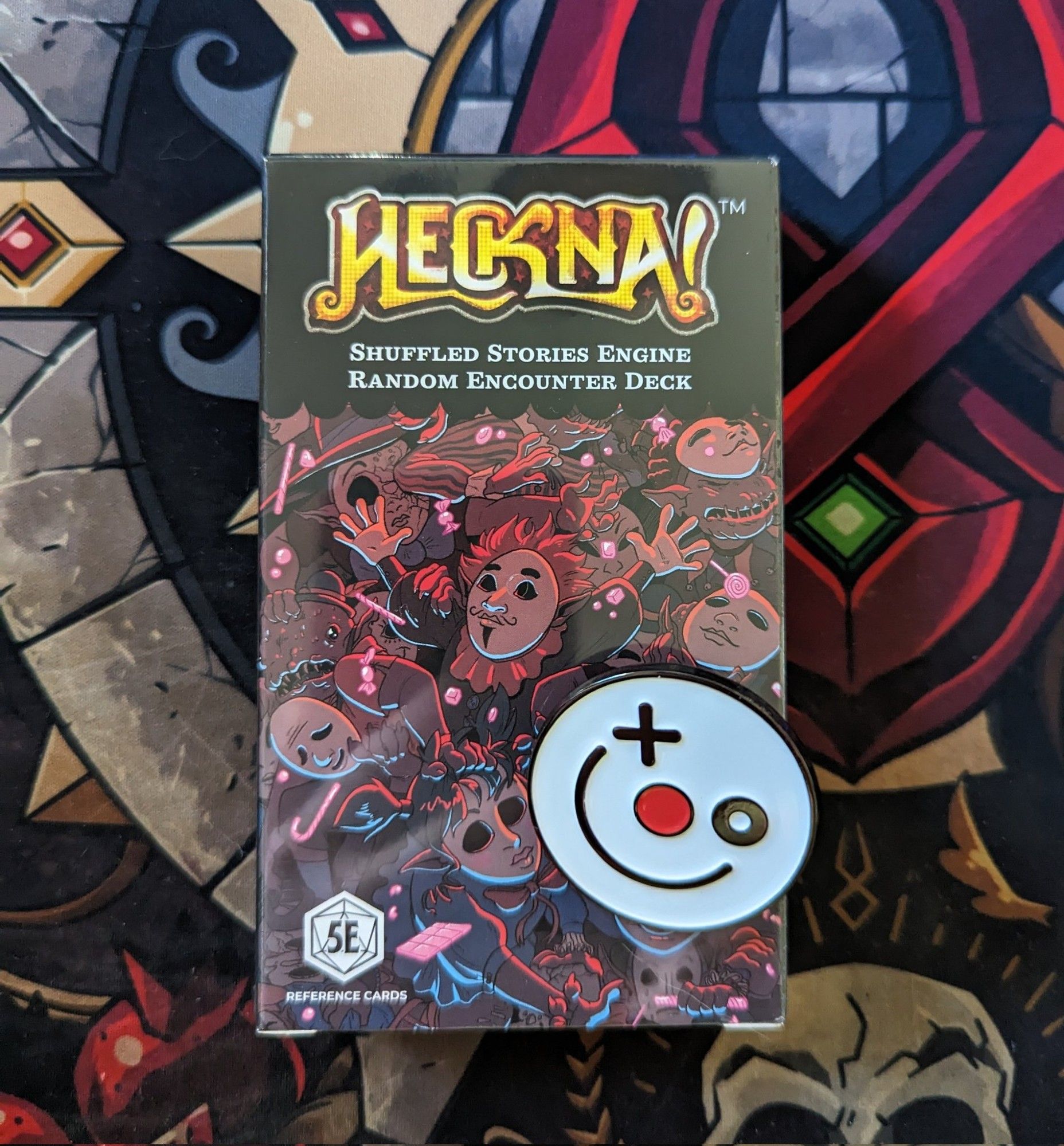 A deck of cards (The Shuffled Stories Engine) with the HECKNA logo, and an enamel pin: A white smiley face with a red nose, and a cross over one of the eyes.