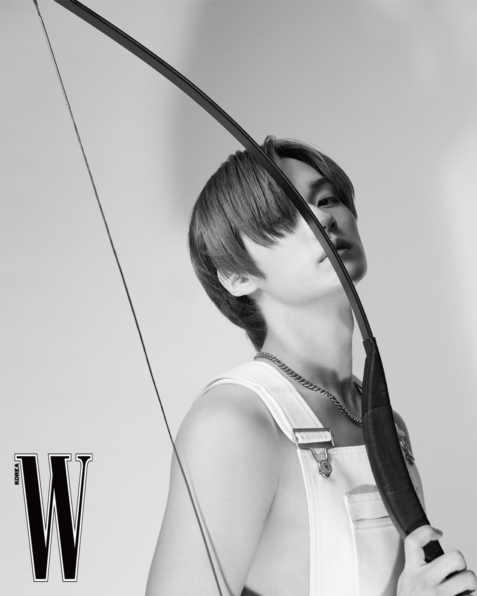 Lee Know in black and white wearing a white overalls and a silver chain in 3/4 stance facing the camera on a light gray background. He is holding a longbow, the arc of it visually cutting his face vertically