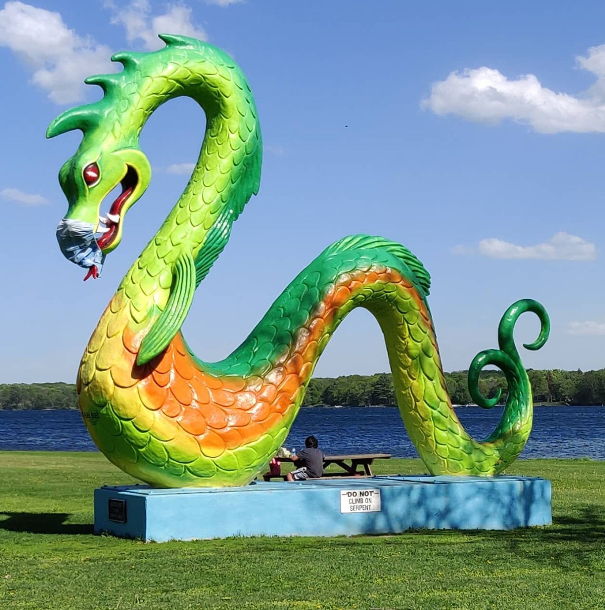 Photo showing large statue of a serpent-style dragon at a community park on the shore of a small lake.  The statue is in shades of green, yellow, and orange, sitting on a blue platform.  There is a "surgical mask" that has been put over the open mouth of the dragon, with the forked tongue punching through the material.