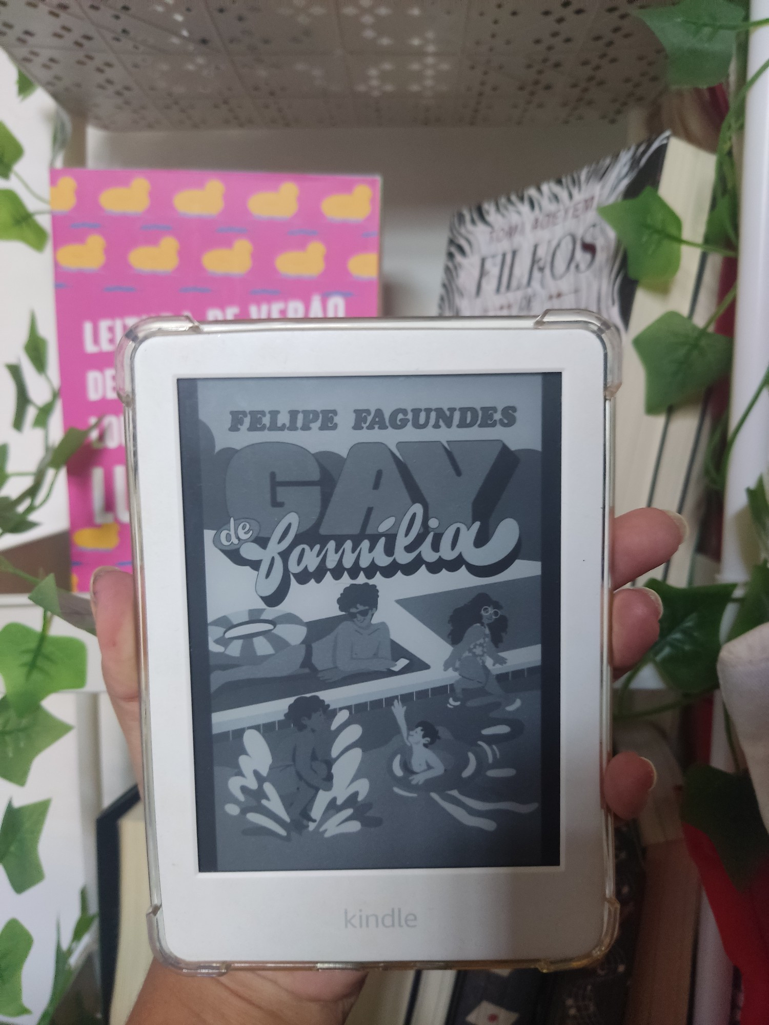 Foto de um kindle com a capa de Gay de Família de Felipe Fagundes. Há um homem na beira da piscina e ele assiste três crianças, dois meninos brincam dentro da água e uma menina está sentada na borda com os pés na água 