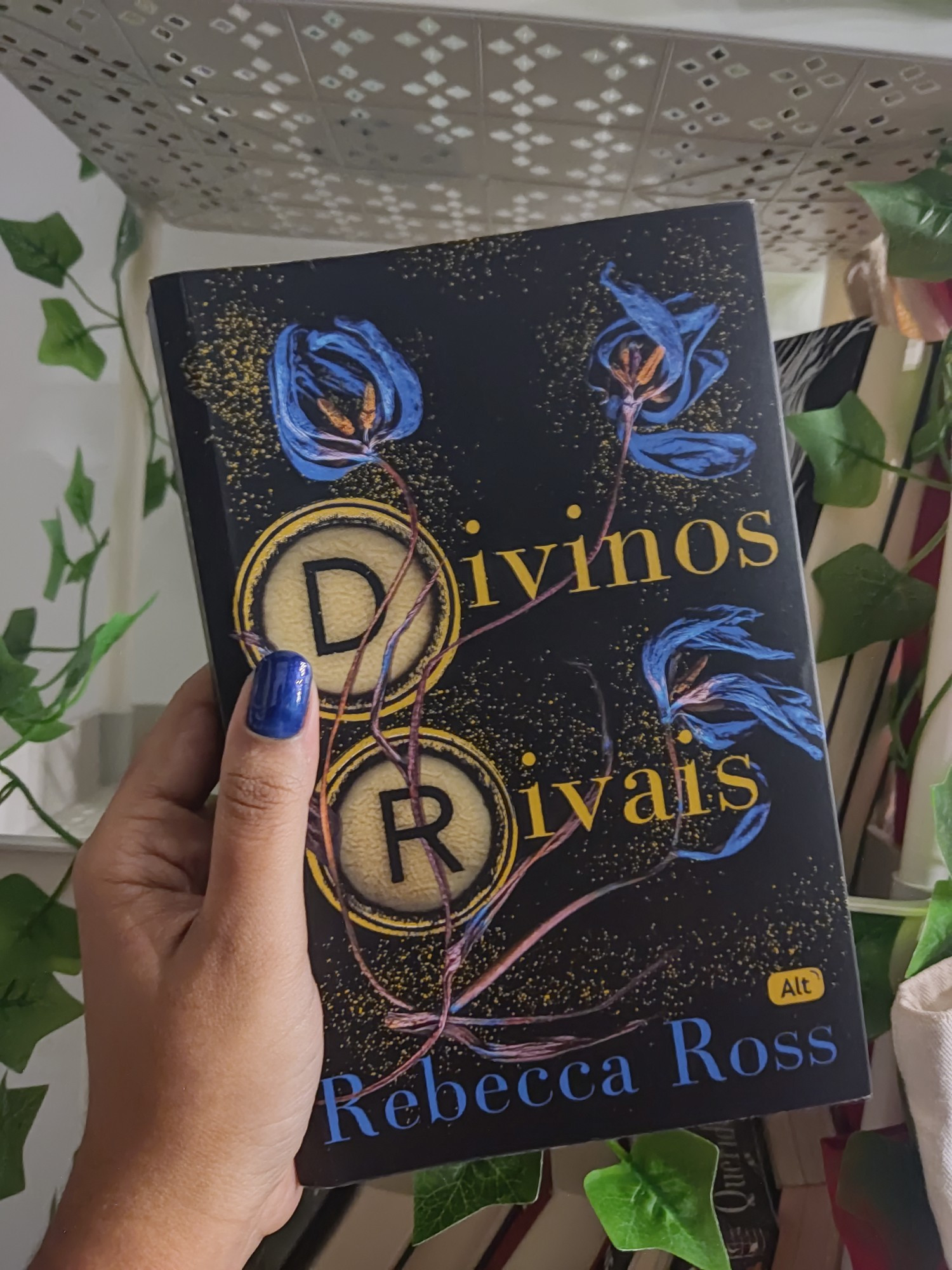 Foto do livro Divinos Rivais de Rebecca Ross. A capa é preta com pontos de luz douradas e há um ramo com três flores azuis 