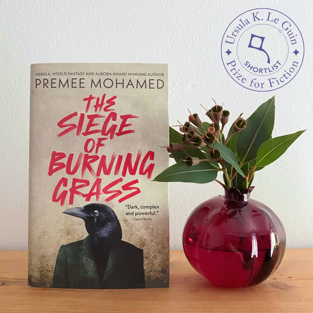 A copy of Premee Mohamed's The Siege of Burning Grass sits on a wooden shelf next to a red glass vase shaped like a pomegranate, holding some green flowers