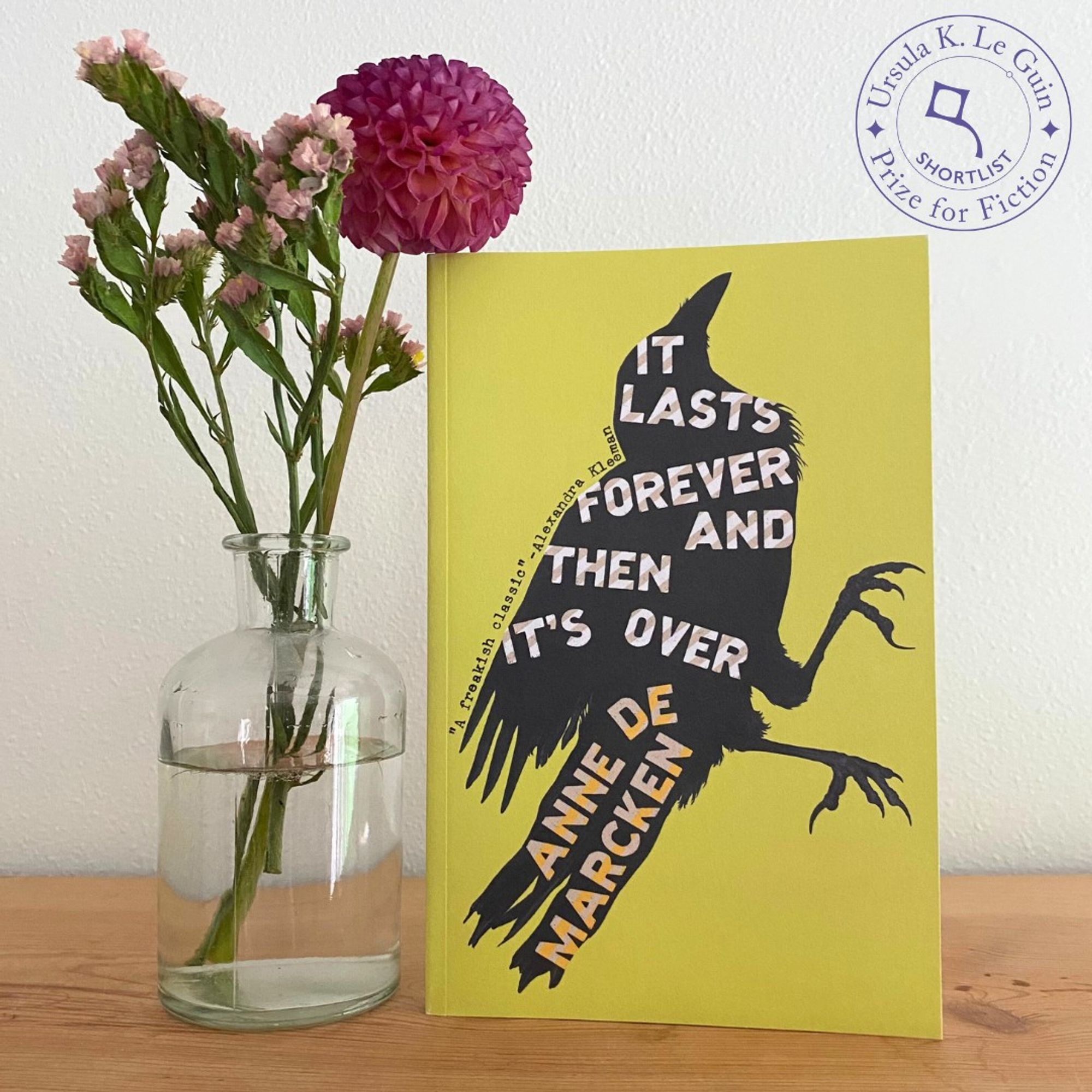 A copy of Anne de Marcken's It Lasts Forever and Then It's Over stands on a wooden shelf next to a glass vase with a round red flower and some small pink flowers