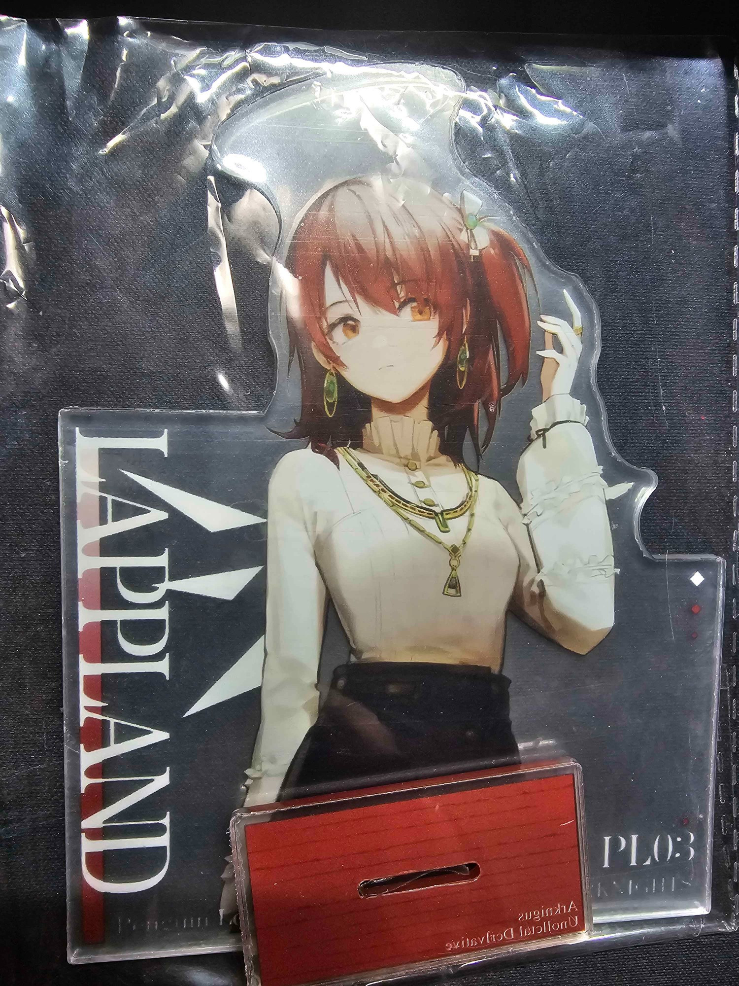 The image shows a misprinted acrylic stand featuring Arknights character Exusiai, but the text humorously labels her as "LAPPLAND." Exusiai, known for her energetic and angelic design, is depicted in a more casual outfit than her usual in-game attire. She has her signature red hair tied up in a ponytail, wearing a white high-neck blouse, dark high-waisted pants, and gold jewelry, including earrings and a necklace. Despite the clear depiction of Exusiai, the bold text to the left of the character reads "LAPPLAND," making this a funny production error for fans familiar with both characters. The stand appears to be part of the "PL03" series, with a base visible below the figure for support.