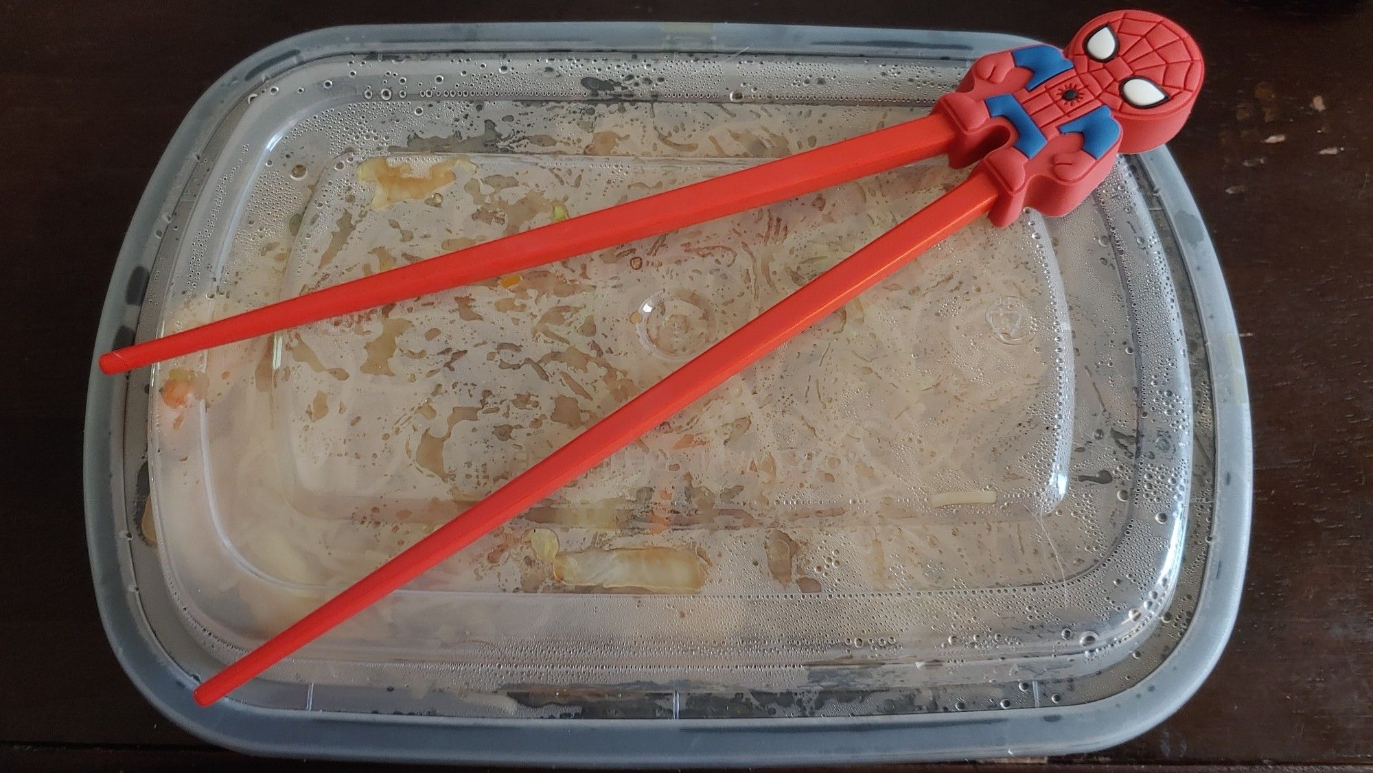 A sealed container of mei fun with red chopsticks on top. The chopsticks are attached to a rubber Spider-Man's legs, making an easier, beginner set of chopsticks.