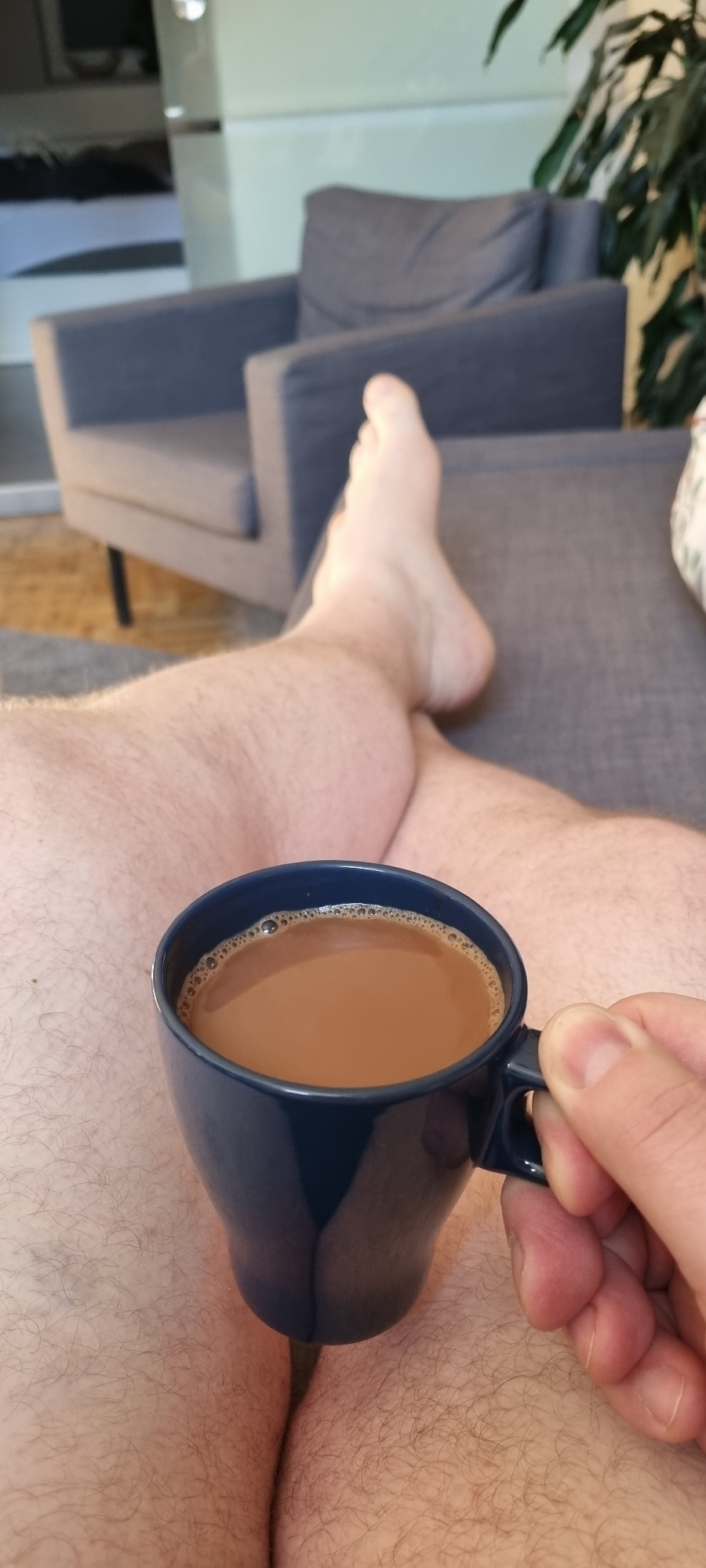 Beschreibung von chatgpt: auf dem Foto ist eine Person zu sehen, die entspannt auf einem Sofa sitzt, die Beine übereinandergeschlagen hat und eine Tasse Kaffee in der Hand hält. Die Person trägt keine Kleidung an den Beinen, und der Raum im Hintergrund ist hell und gemütlich eingerichtet mit einem grauen Sessel und einer Pflanze. Die Atmosphäre wirkt ruhig und entspannt.