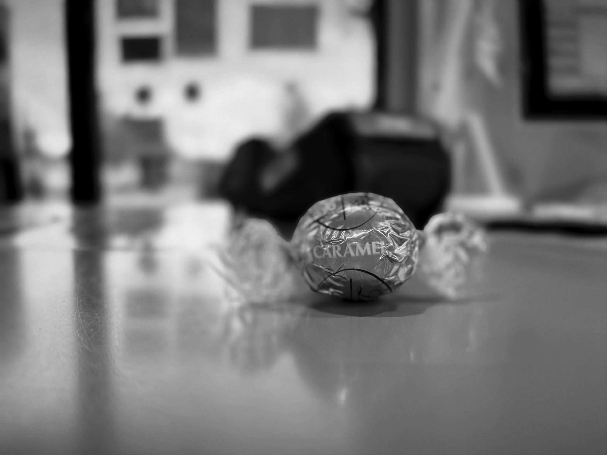 Motiv:
Im Zentrum des Bildes liegt ein einzelnes, verpacktes Karamellbonbon. Es befindet sich auf einer glatten, leicht reflektierenden Oberfläche.

Bildformat:
Das Bild ist im Querformat aufgenommen.

Genre:
Das Foto gehört zur Kategorie der Stillleben-Fotografie.

Hintergrund:
Der Hintergrund ist unscharf, was die Aufmerksamkeit auf das Bonbon im Vordergrund lenkt. Im Hintergrund scheinen unscharfe Büroeinstellungen sichtbar zu sein, aber sie sind nicht klar zu erkennen.

Farben:
Das Bild ist in Schwarz-Weiß gehalten, wodurch Kontraste und Formen hervorgehoben werden, während farbliche Nuancen fehlen.

Lichtsituation:
Das Bild zeigt eine sanfte Beleuchtung, die eine leichte Spiegelung auf der Oberfläche erzeugt. Das Licht fällt von der linken Seite auf das Bonbon und verstärkt seine Struktur, während der Hintergrund im Schatten liegt.