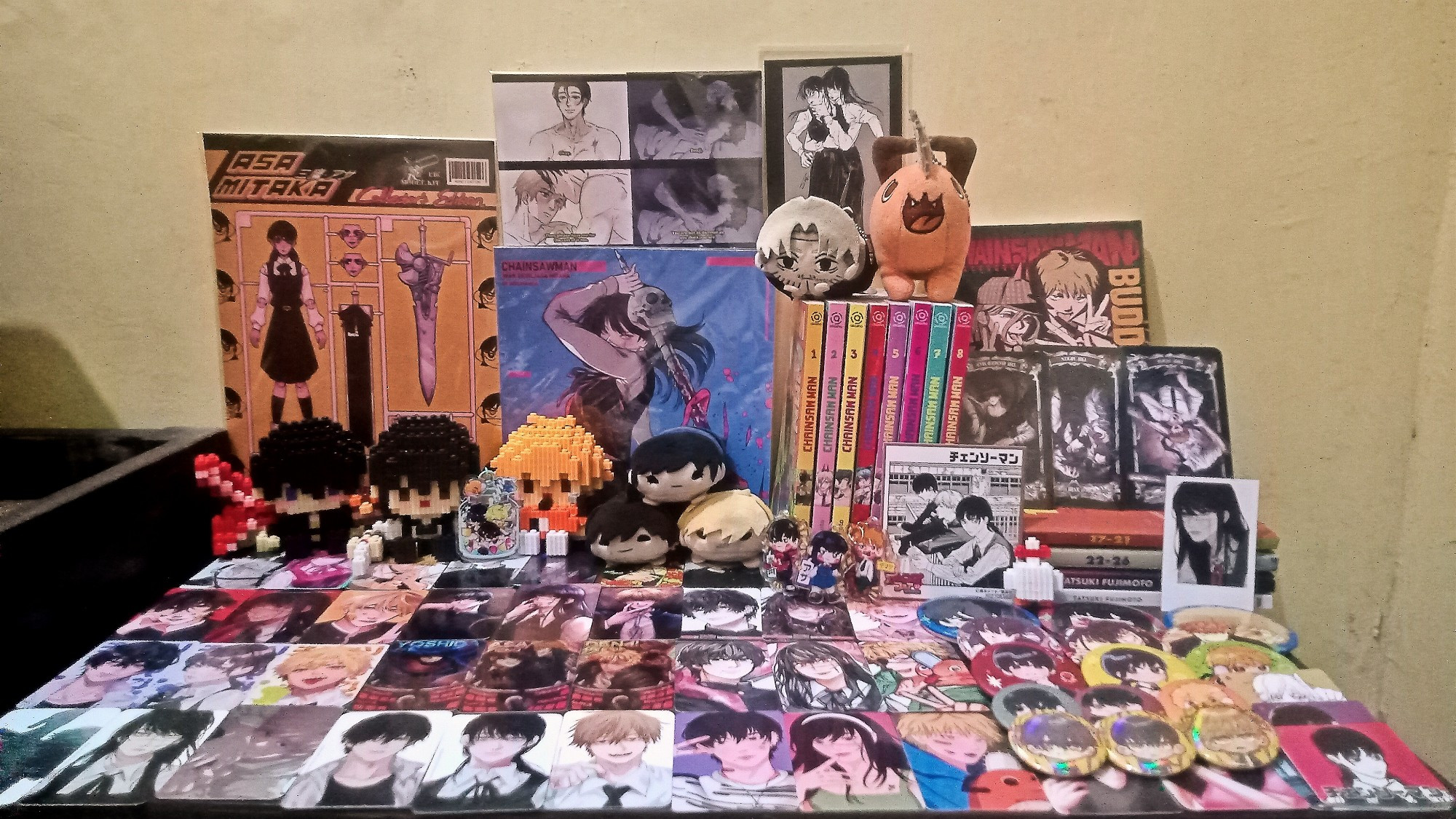 My yoshidenasa merchs and fanmerchs shrine, consists of: Chainsawman mangas currently available in Indonesia, Fujimoto's oneshots, Buddy Stories novel, posters, silly yoshiden and asayoru prints, the official shikishi and noru charas, FA photocards and badges, and unofficial bricks.