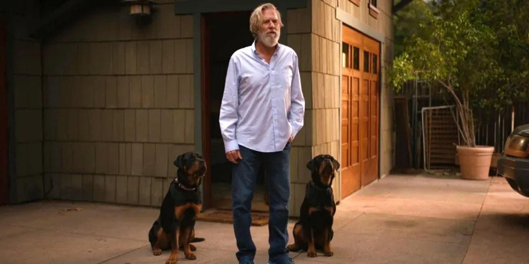 Still from The Old Man, Jeff Bridges standing outside a house with a rottweiler on either side of him, both sitting obediently, always ready to kill yet another highly-trained assassin.