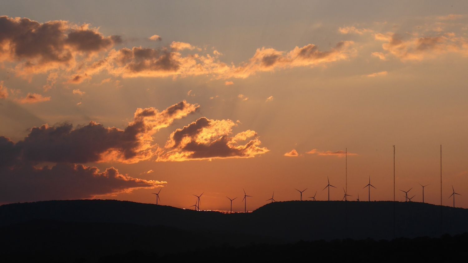 Several hills form the black horizon line about 1/4 the way up the photo. Above this the sky is orange fading to steel blue at the top of the photo. Several clouds above the horizon add red and yellow shades to the sunset color, and rays of reflection are faint but visible, radiating out of the center cloud. Above the horizon line and just below the sunset are a series of wind turbines. There are apparently terrifying to Republicans, for some reason.