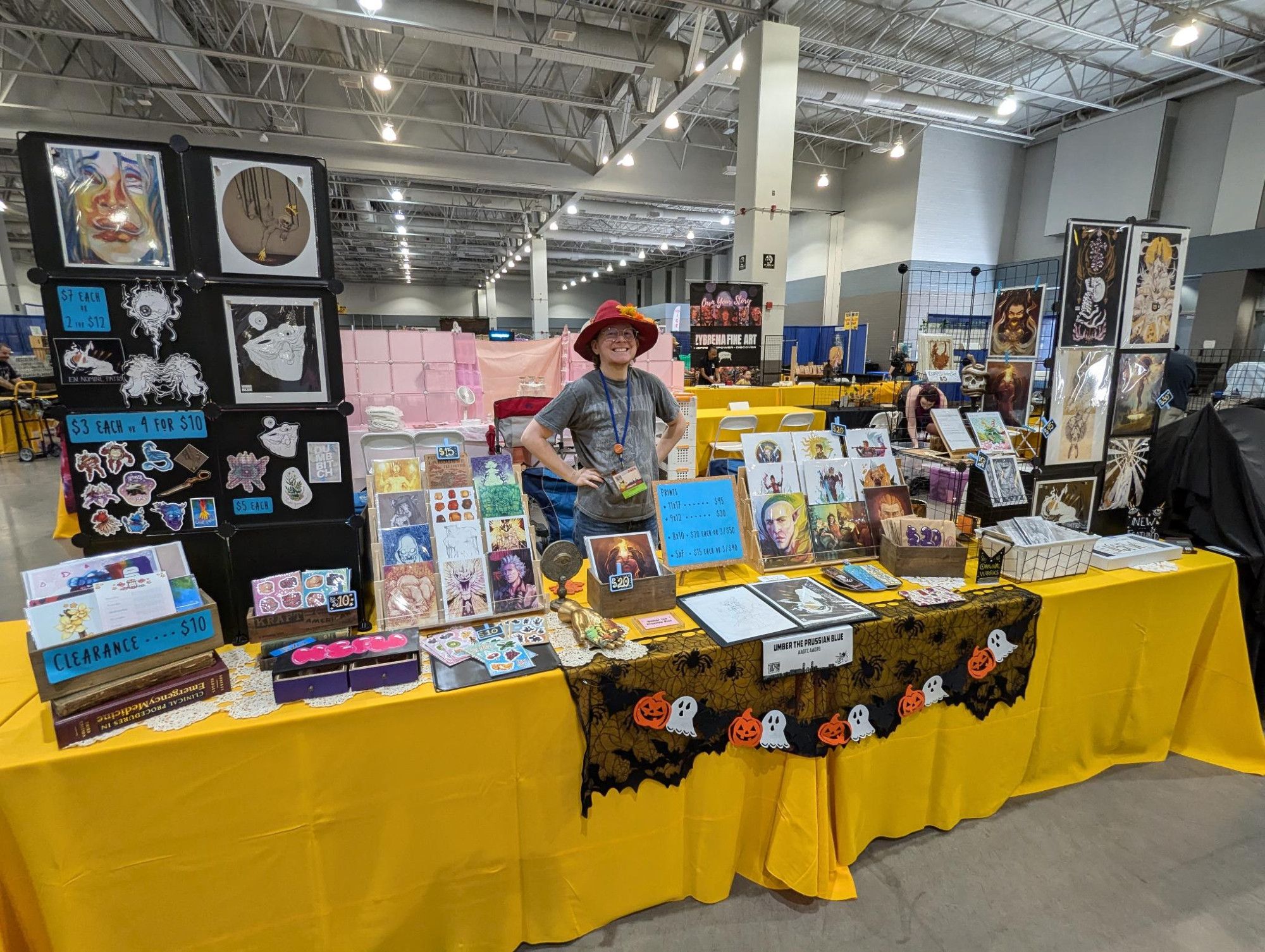 A photo of me standing at my table during set up day. I have two tables with a wide selection of different goods like prints and stickers. I'm also wearing a rad pointy hat.