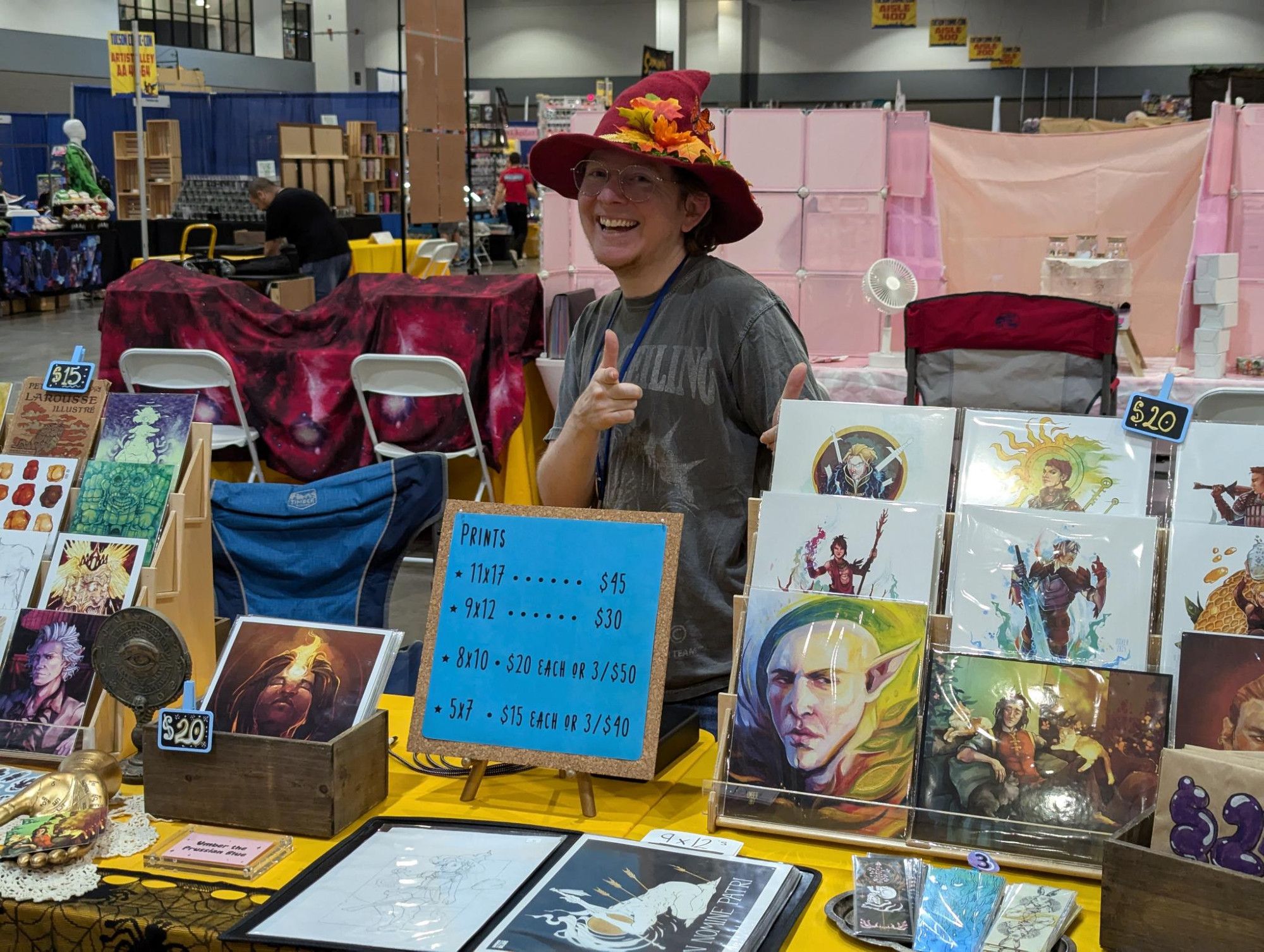 A close up of me in my pointy hat giving very bisexual finger guns. My dragon age prints are featured in this photo